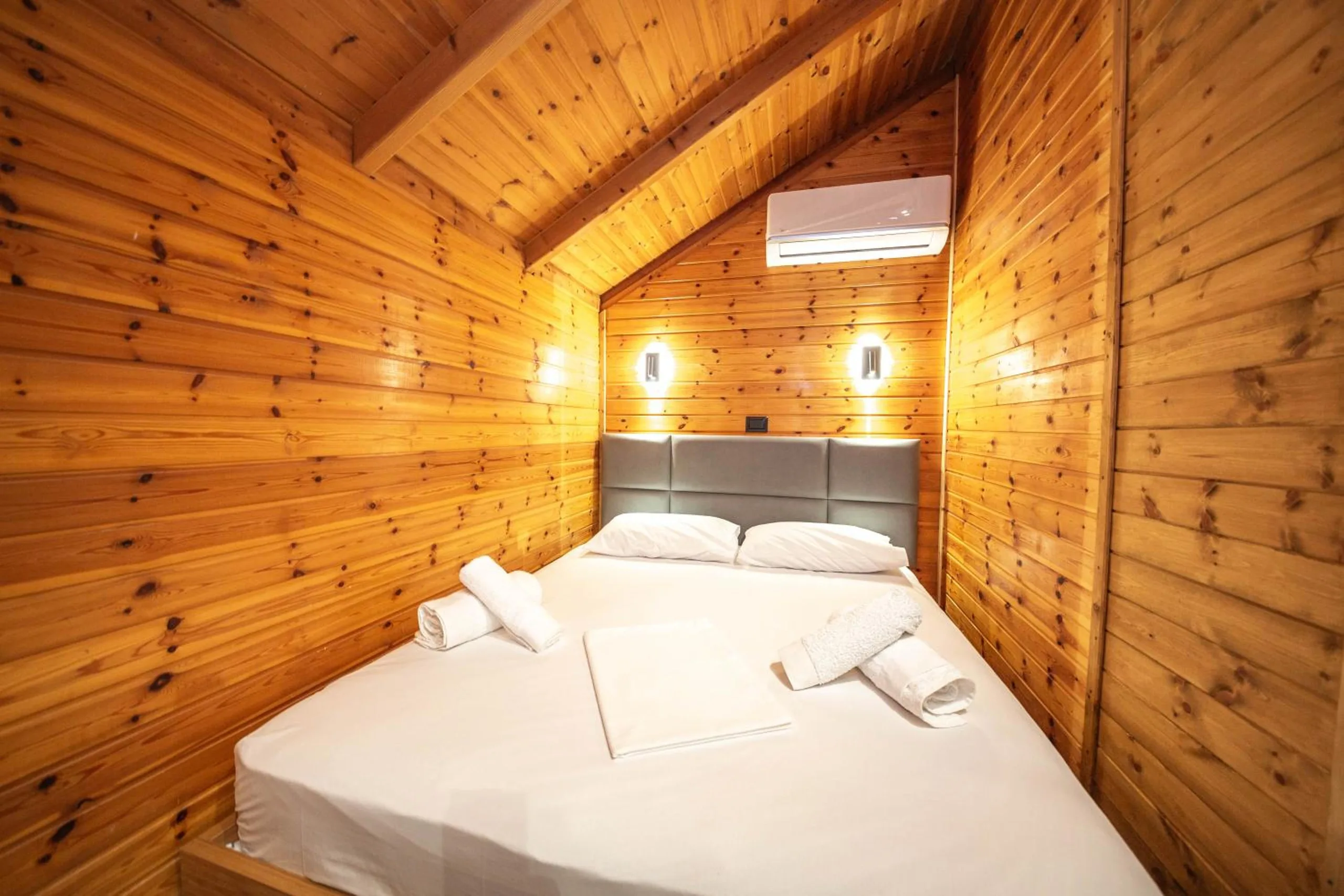 A queen bed with a grey headboard between two wall lights, set against knotty-pine boarding under an air-conditioning unit