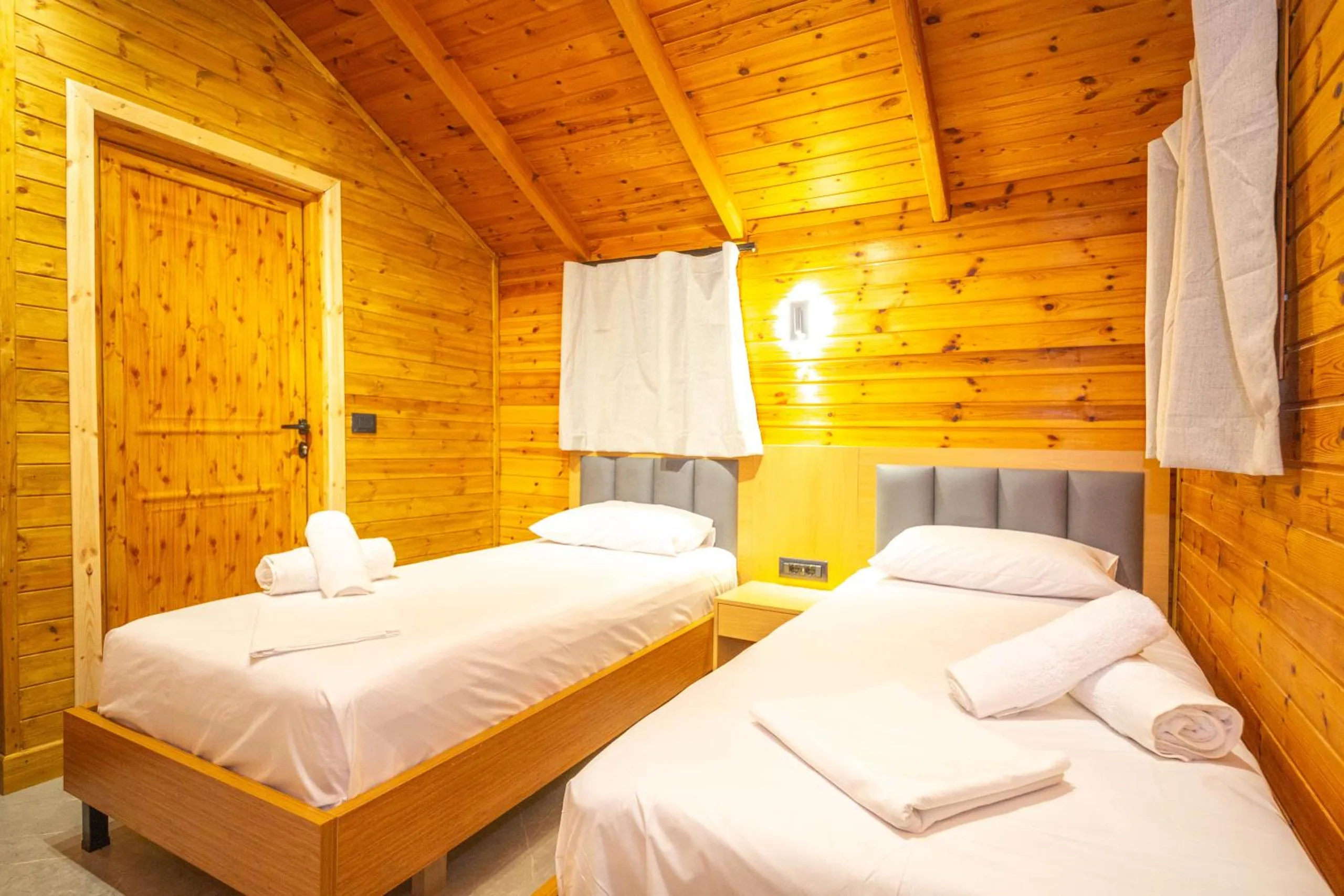 Two single beds on oak frames with grey headboards in a pine-boarded room, a pine door and a curtained window behind