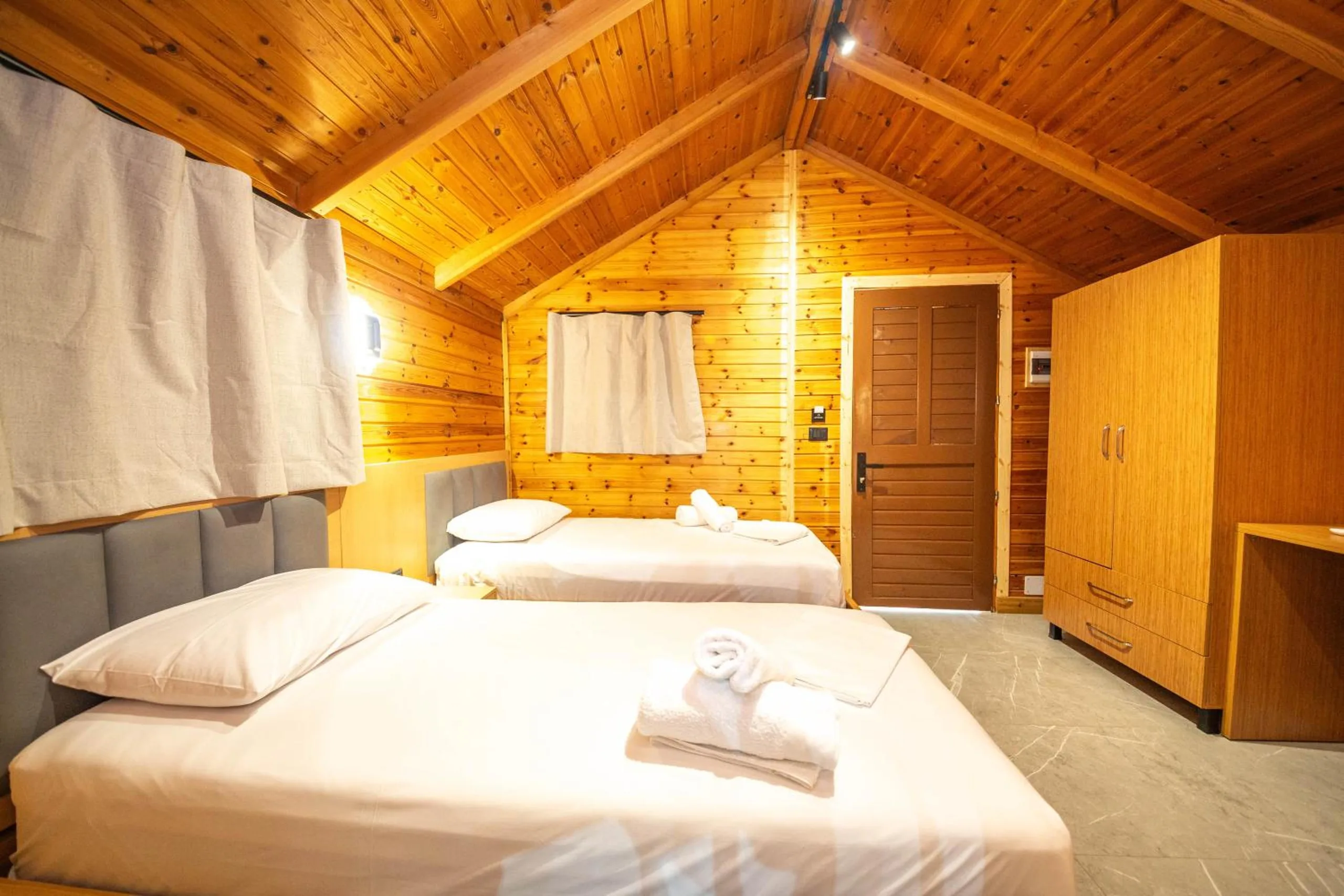 Two single beds with rolled white towels in a pine-boarded bungalow, a tall pine wardrobe and a slatted brown door