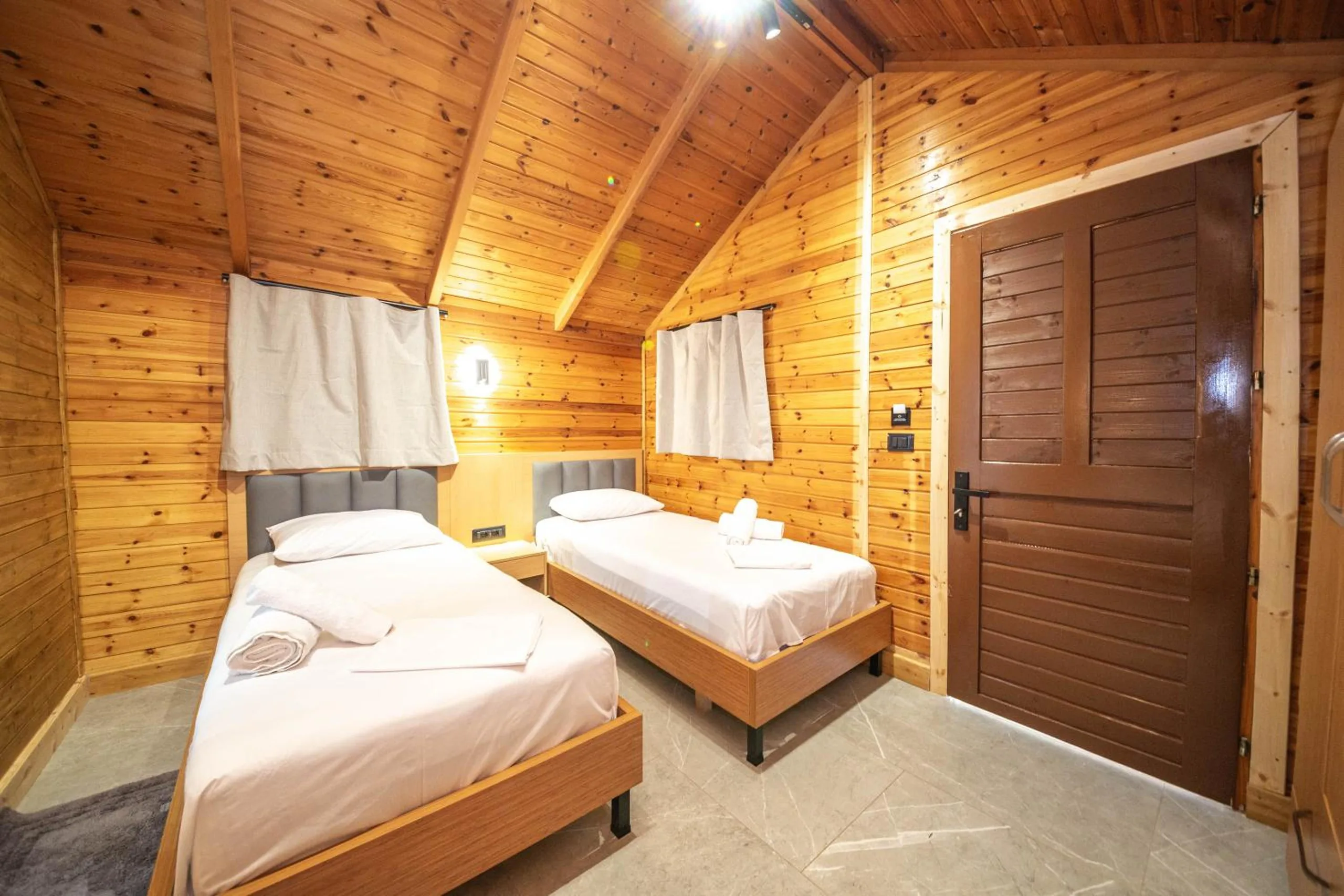 Single beds in a pine-boarded bungalow beside a dark slatted door, a curtained window above