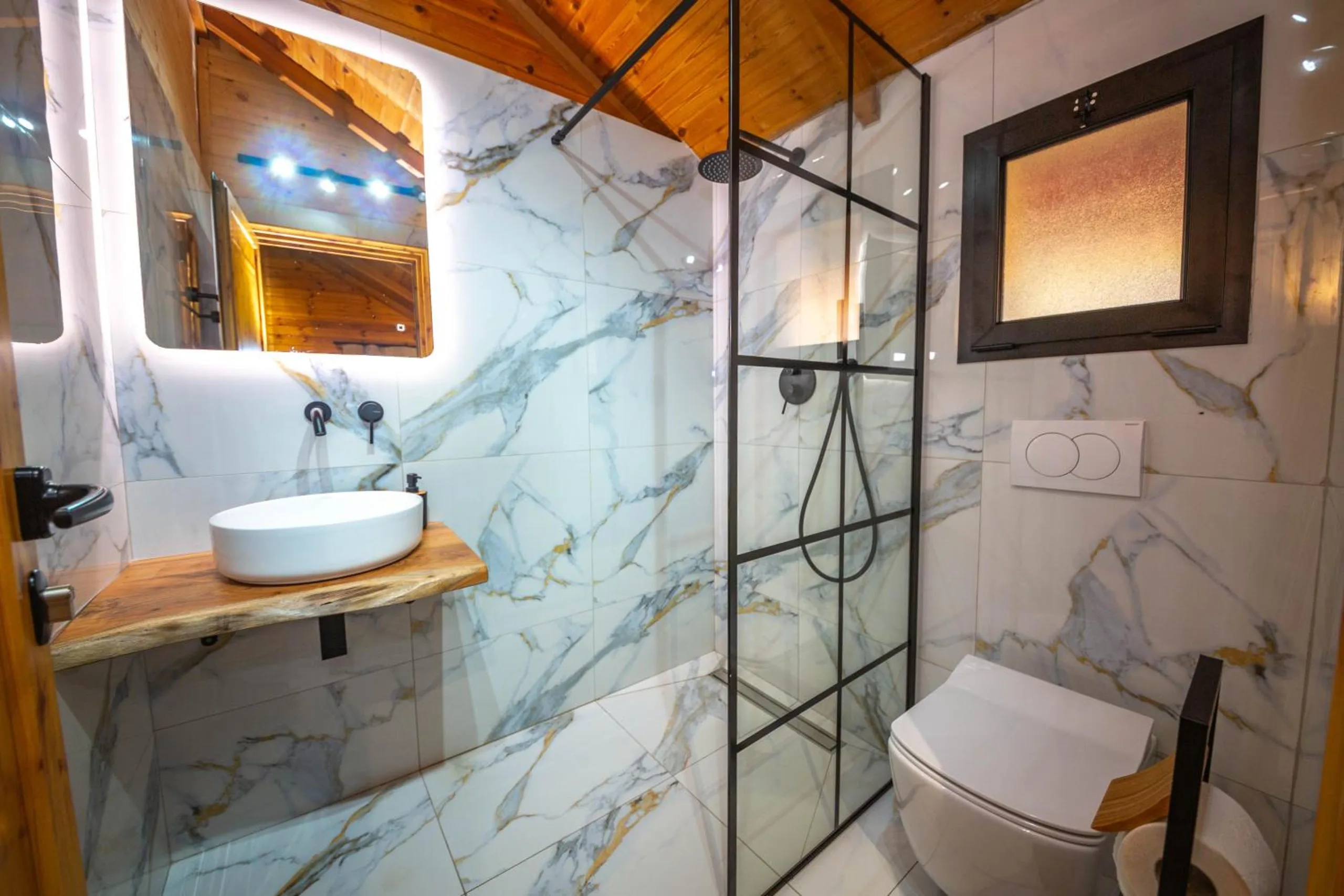 A shower room in white marble-effect tile with a black-framed glass shower screen, a round basin on a live-edge wooden shelf and a backlit mirror