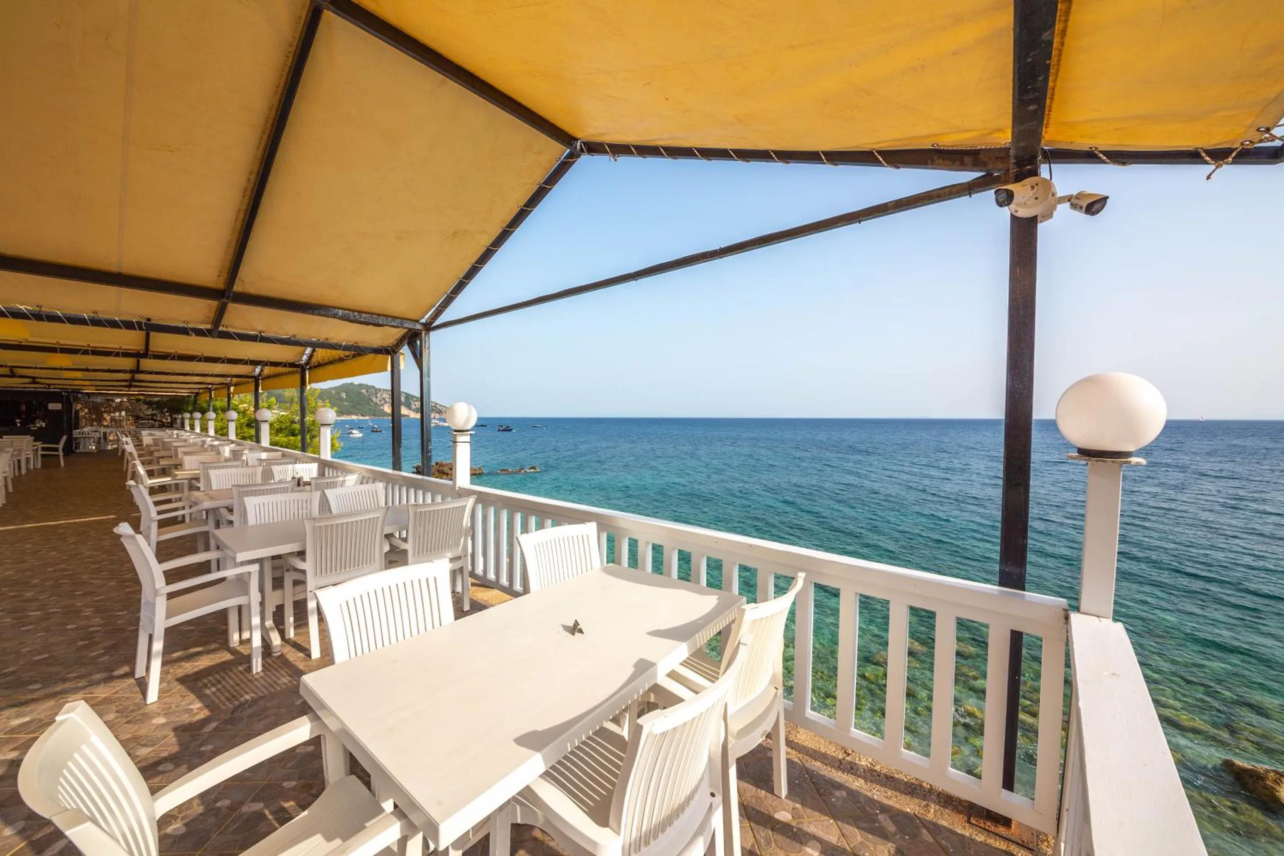 White tables and chairs on the covered restaurant deck, a globe lamp on the rail and the open Ionian beyond, boats moored off the headland