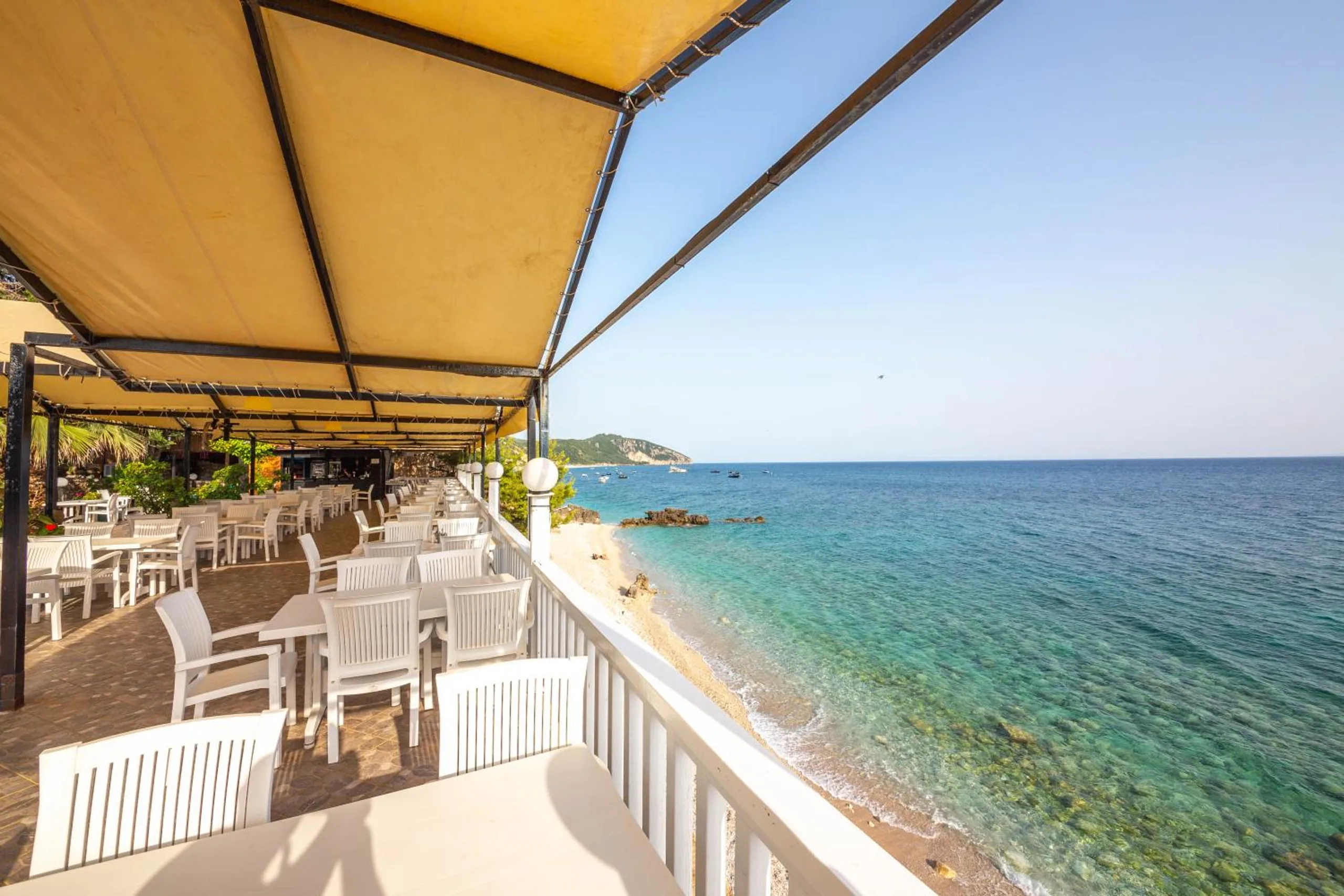 The restaurant deck at Lodges Shën Nikolla under a yellow canvas awning, white tables along a white rail, with the shingle of Dhërmi beach and turquoise water directly below
