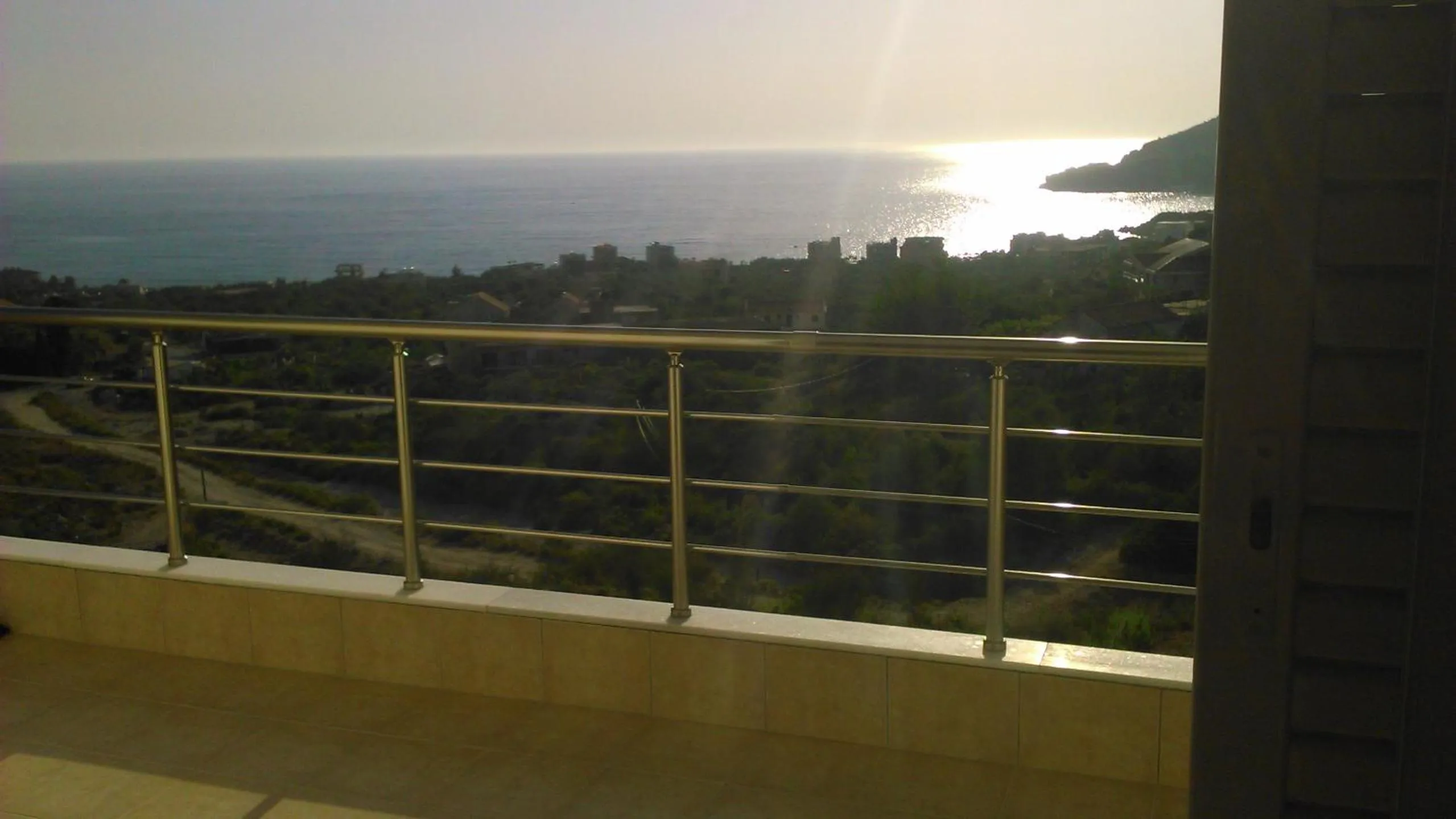 A balcony in low evening sun, its steel rail in silhouette and the sun's reflection on the sea beyond the olive groves
