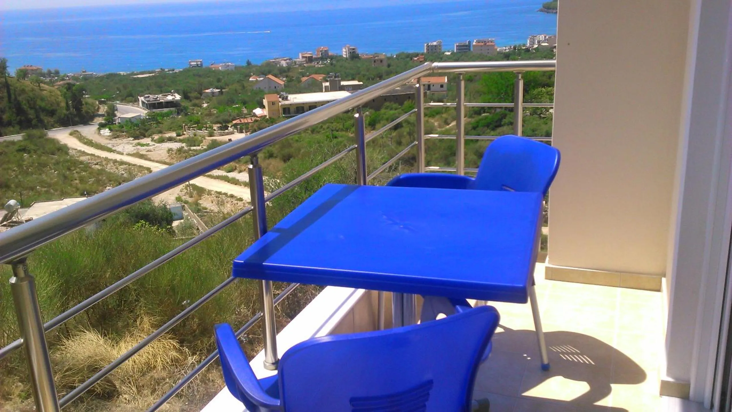 A balcony corner with a blue plastic table and two blue chairs behind a steel rail, the hillside and the sea beyond