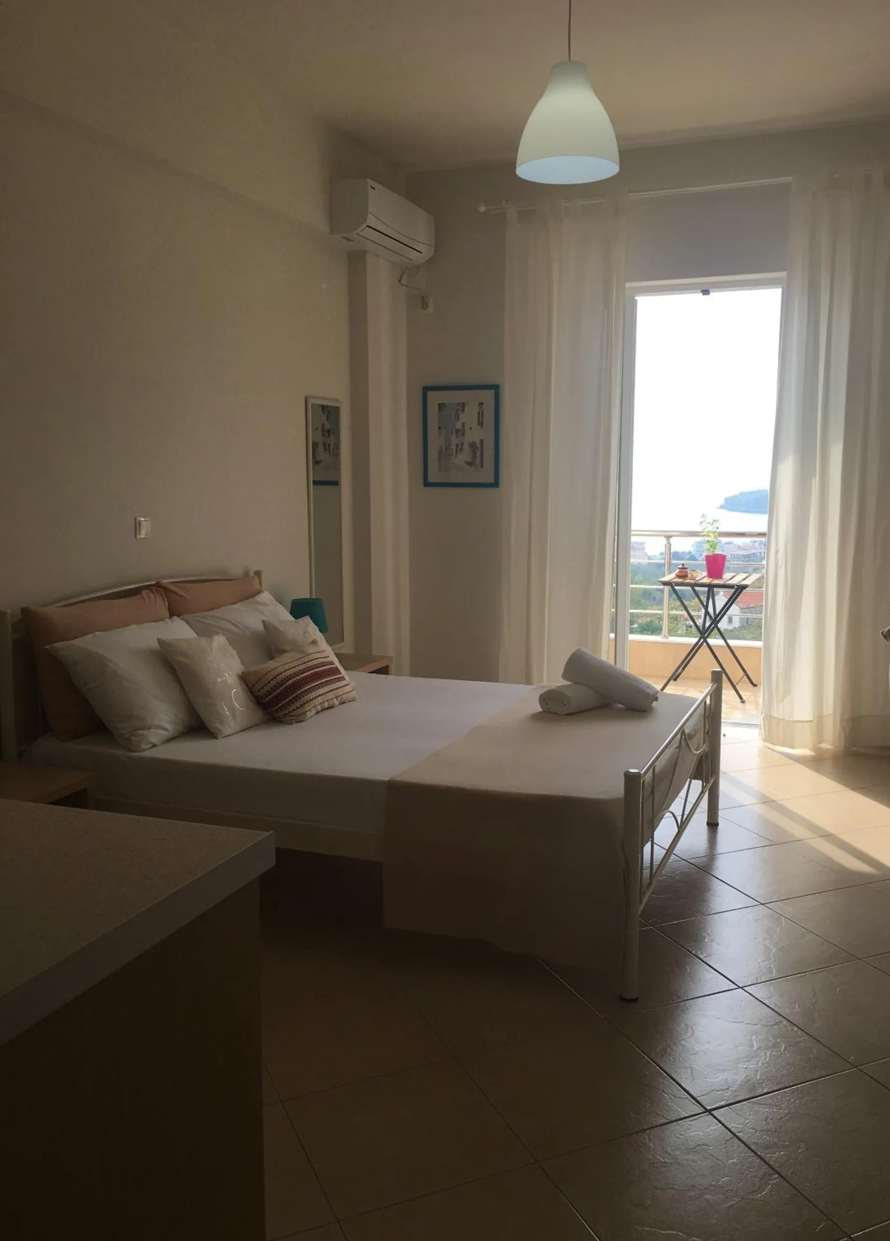 A double room with a pendant lamp and air conditioning unit, sheer curtains drawn back from a balcony door with a small table outside