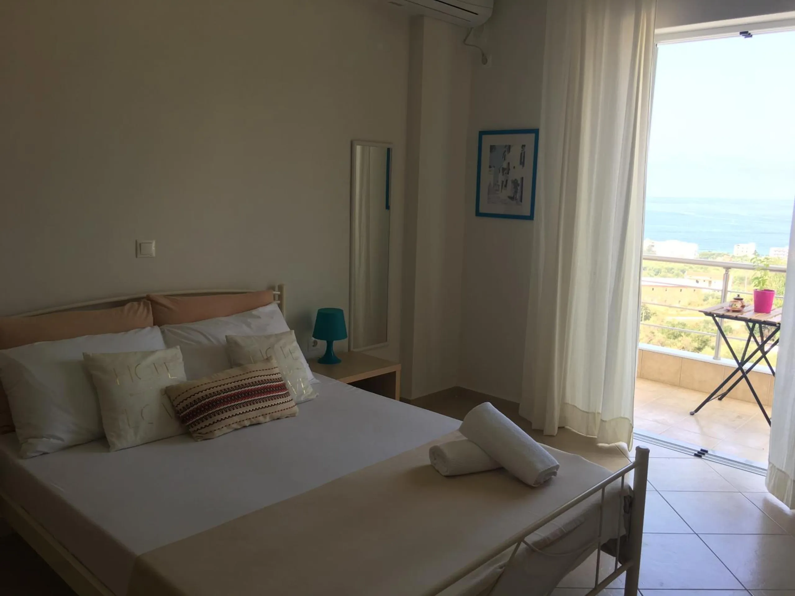 A double bed with striped cushions and rolled towels, a turquoise lamp, and an open balcony door with the sea beyond