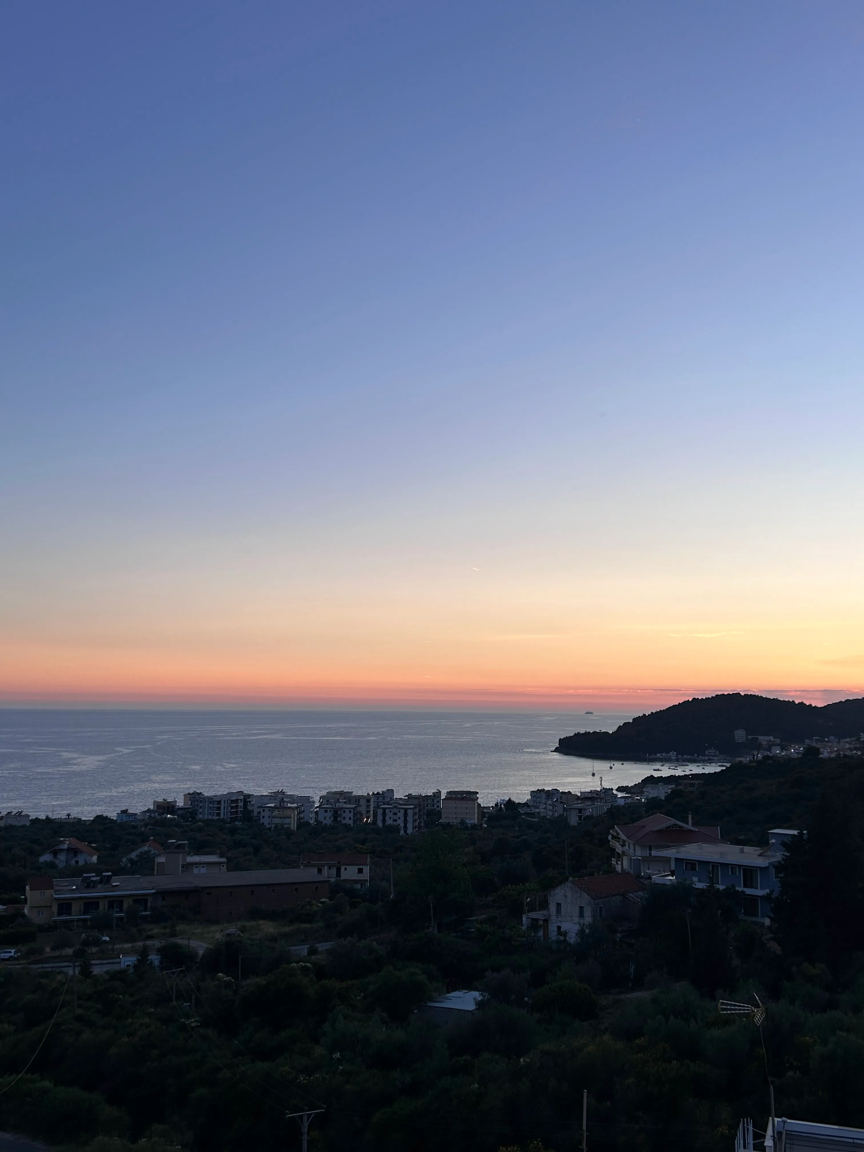 Dusk from the hill above Potam: the sea pale under an orange band of sky, the headland dark on the right and apartment blocks along the shore below the olive groves