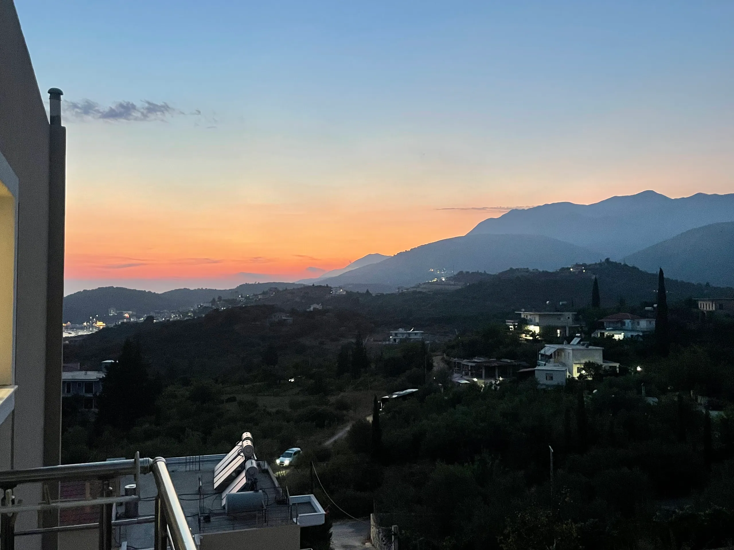 After sunset: an orange band behind the mountains, the lights of the coast coming on to the left, and olive groves going dark below the balcony