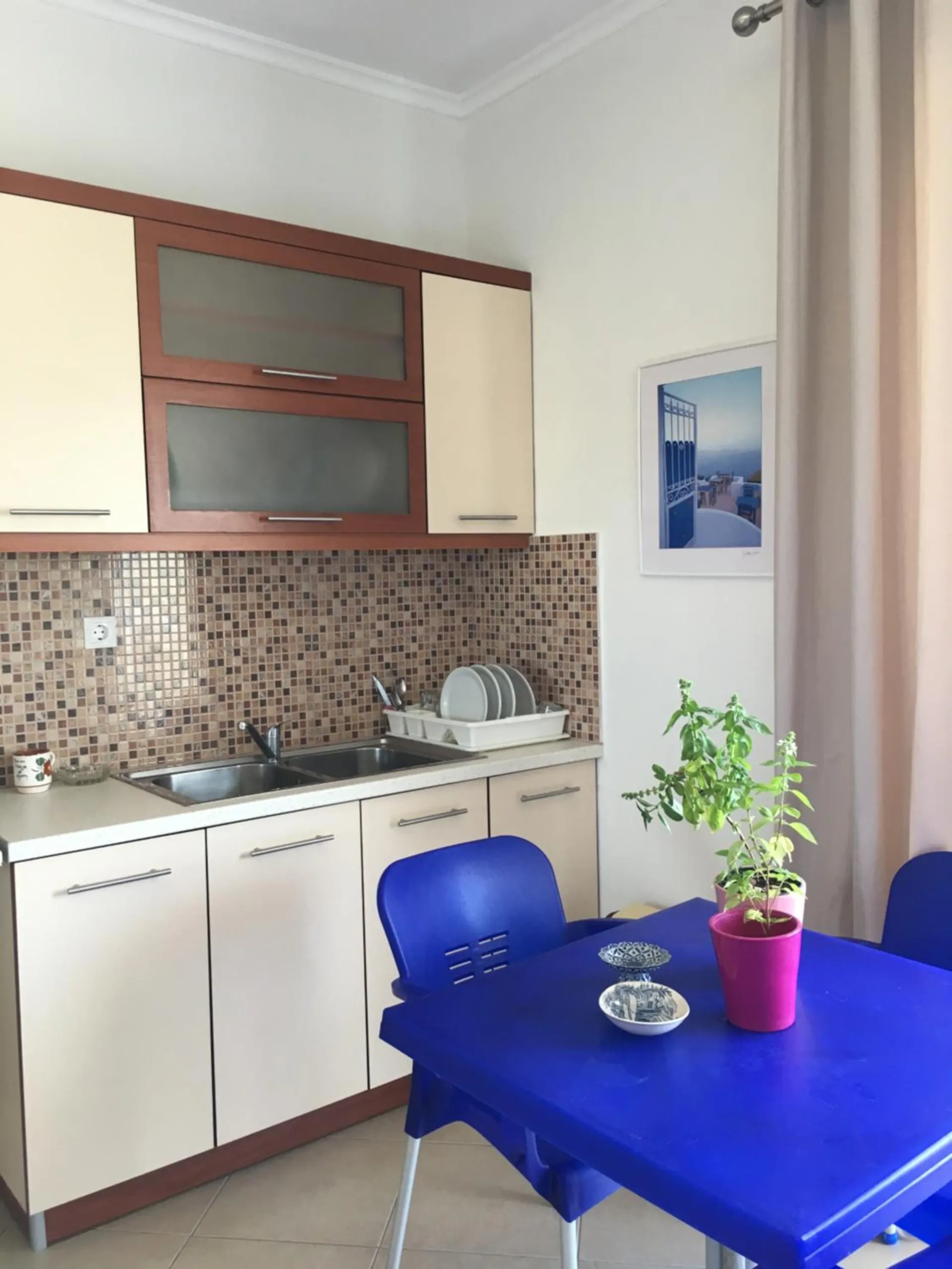 A kitchen corner with cream cupboards, a mosaic splashback, a dish rack, and a blue table with a potted basil plant