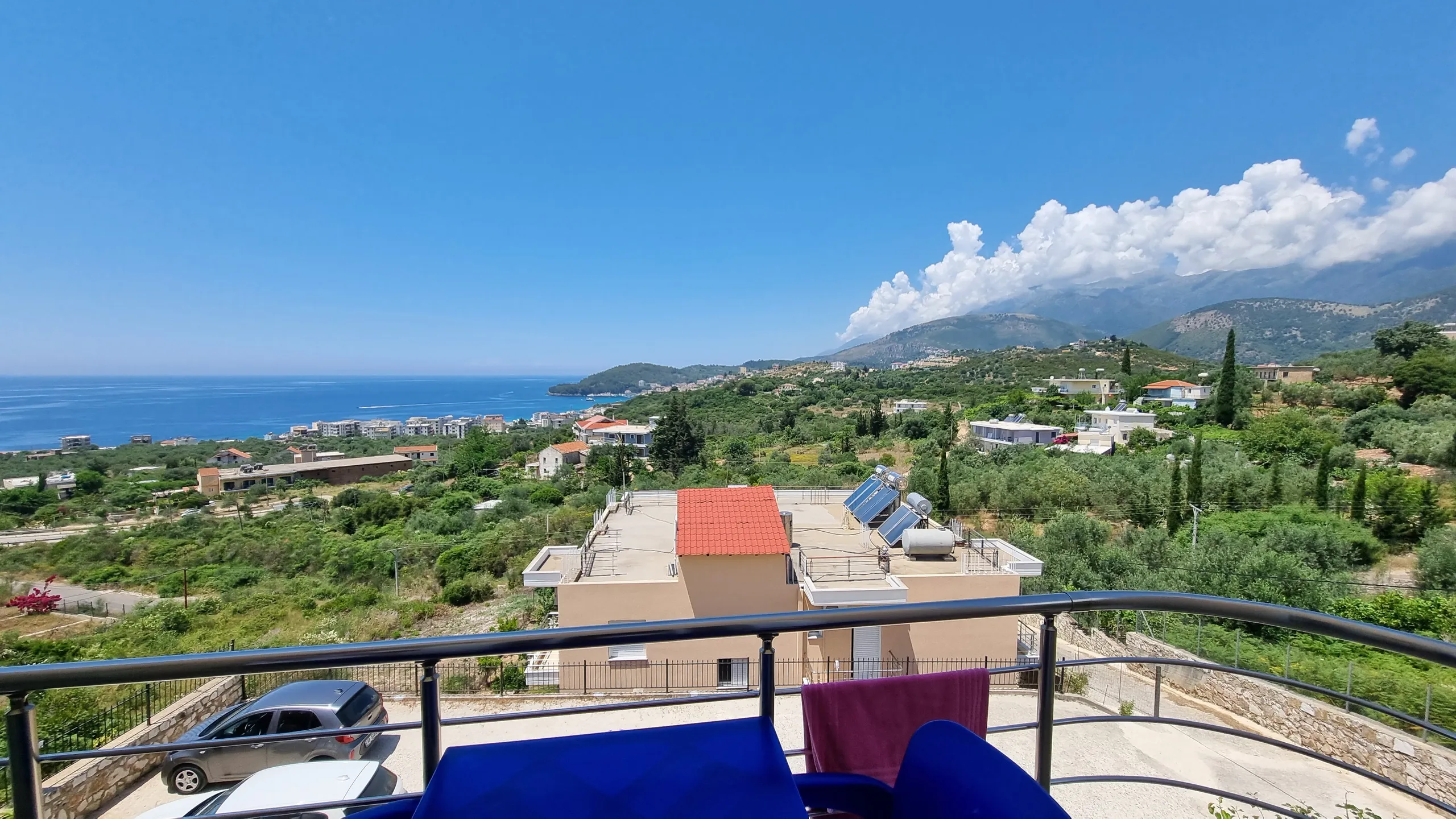The view from a balcony with an ultramarine plastic table and chairs: a red-roofed neighbour, olive groves, the blue bay on the left and mountains under cloud on the right
