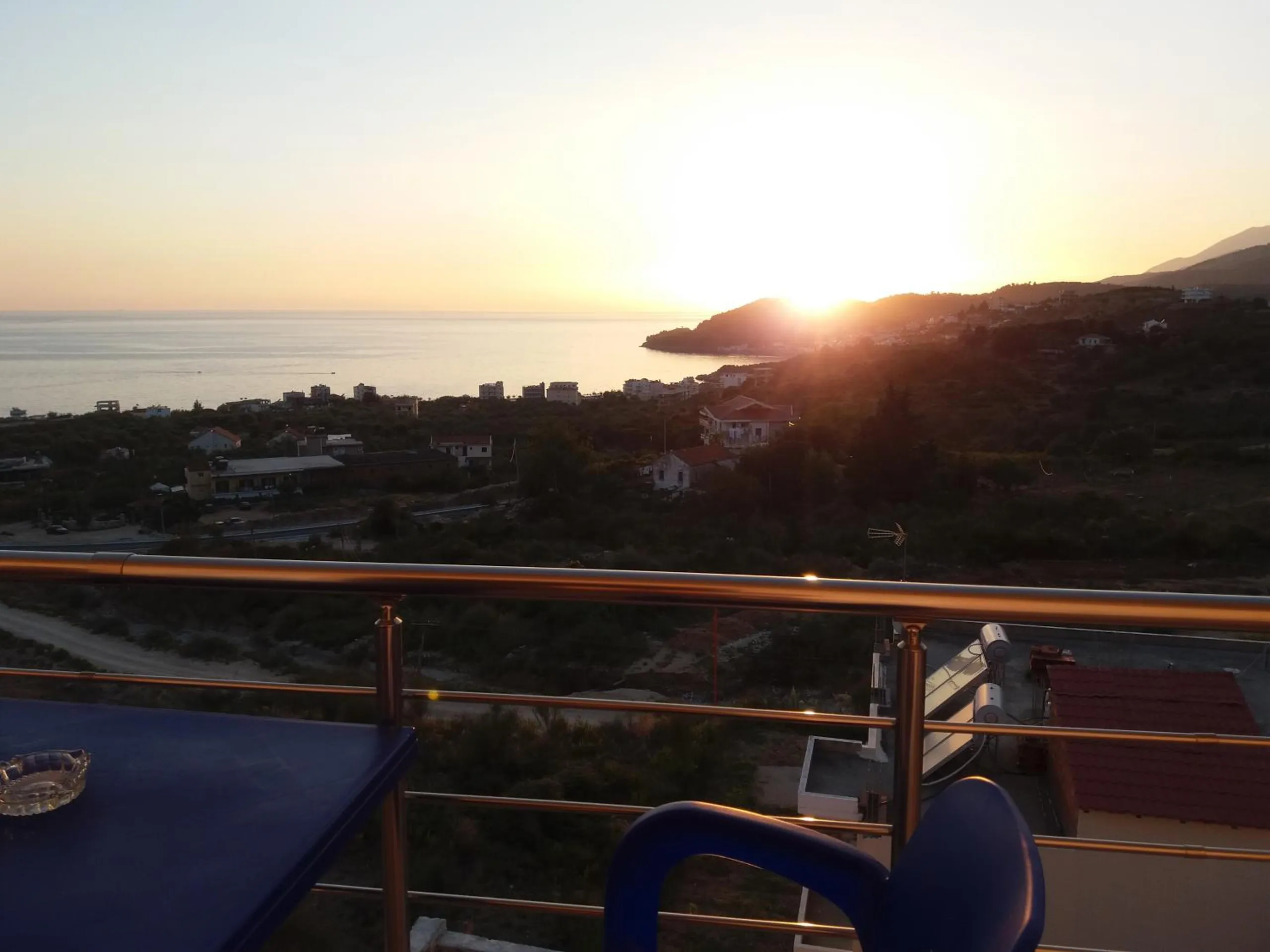 The sun setting behind a headland over the bay, seen past a stainless steel balcony rail, a blue table and the back of a blue chair