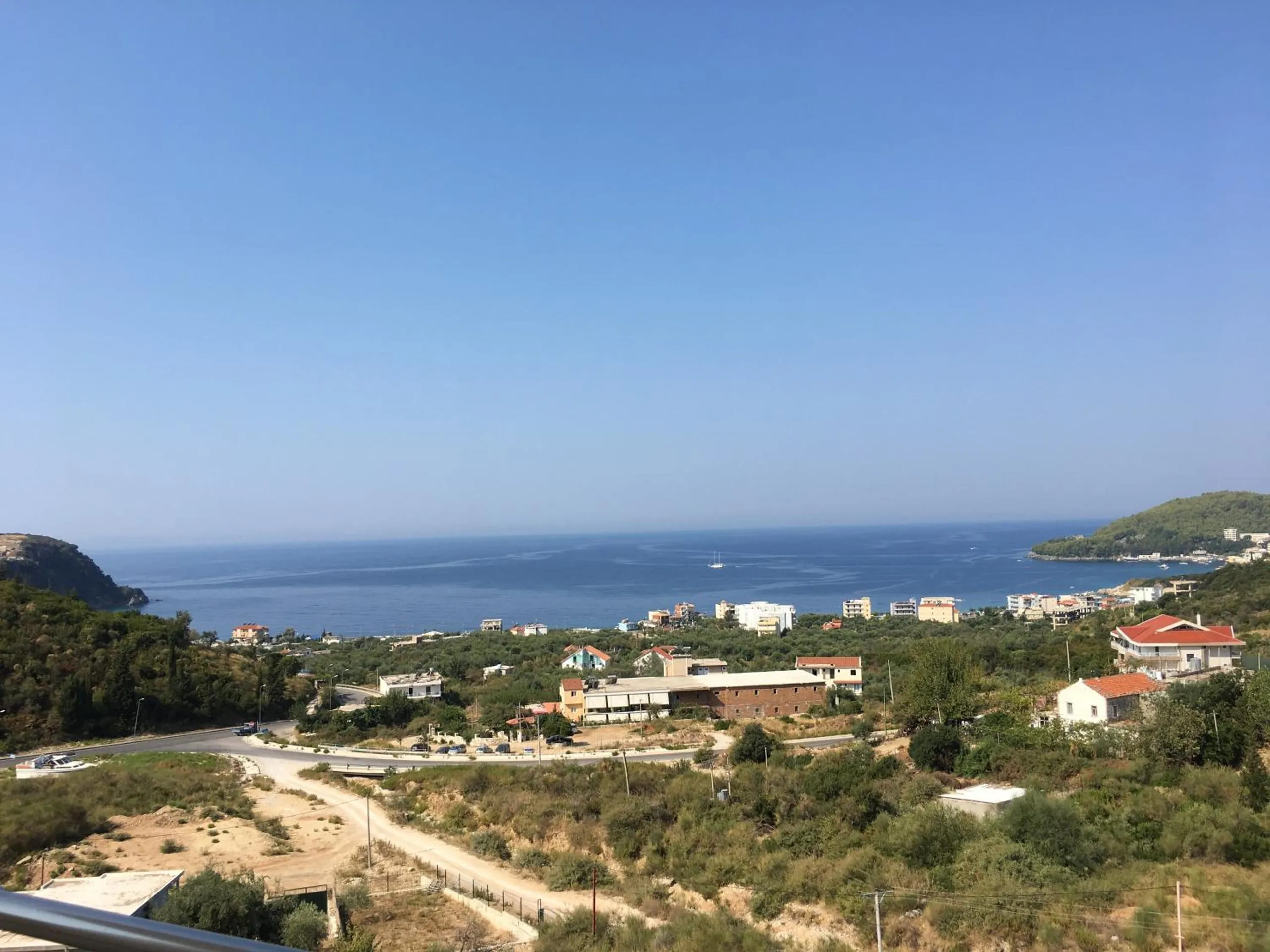 A clear morning over the bay: the coast road curving below, olive groves and houses, two headlands and a sailing boat on flat blue water