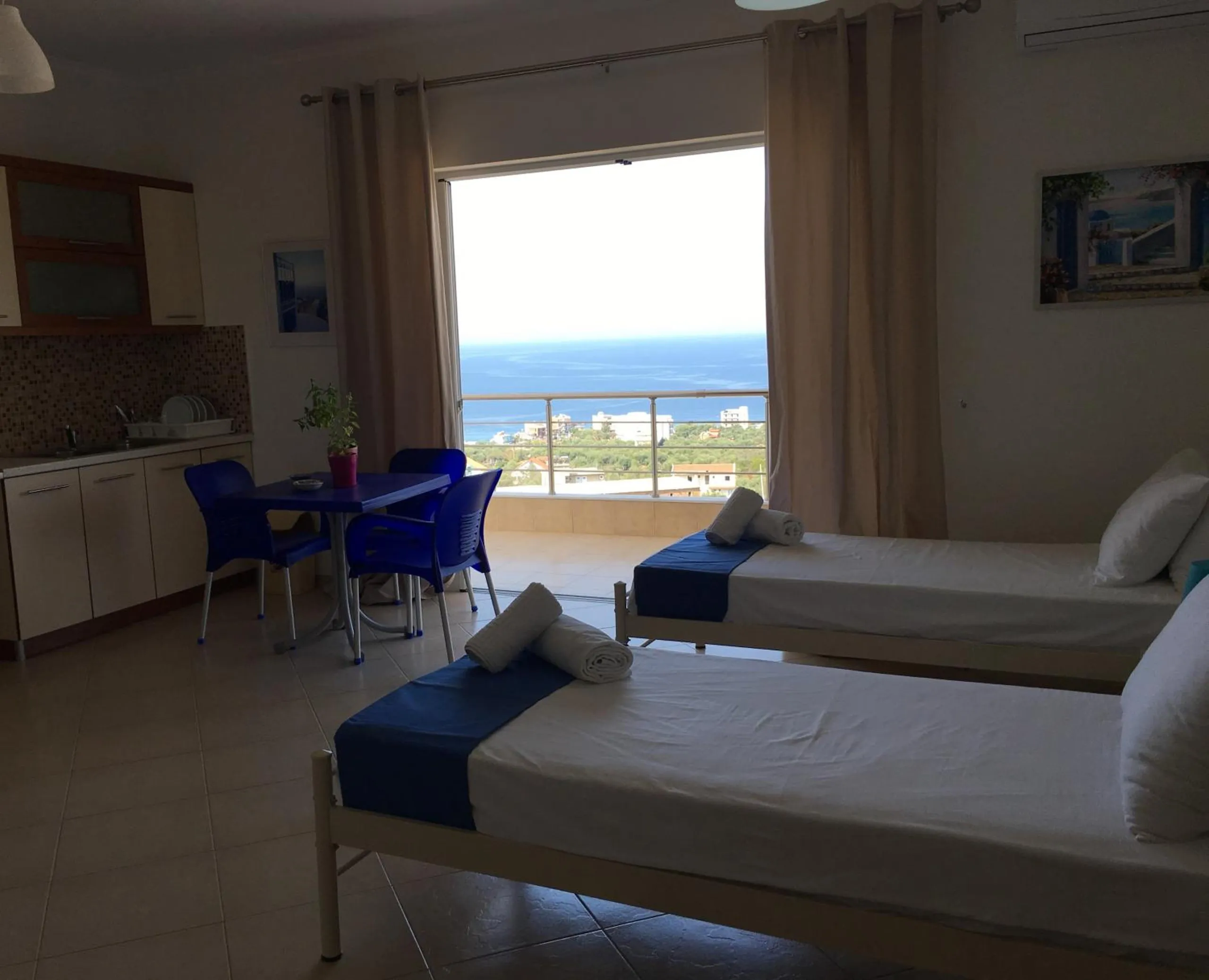 Two single beds with blue throws, a kitchen run and a blue table and chairs, and a wide window onto the sea