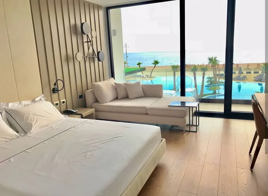 A guest room at Miamar with a king bed, a cream sofa and a full-height window onto the pool and the Ionian Sea