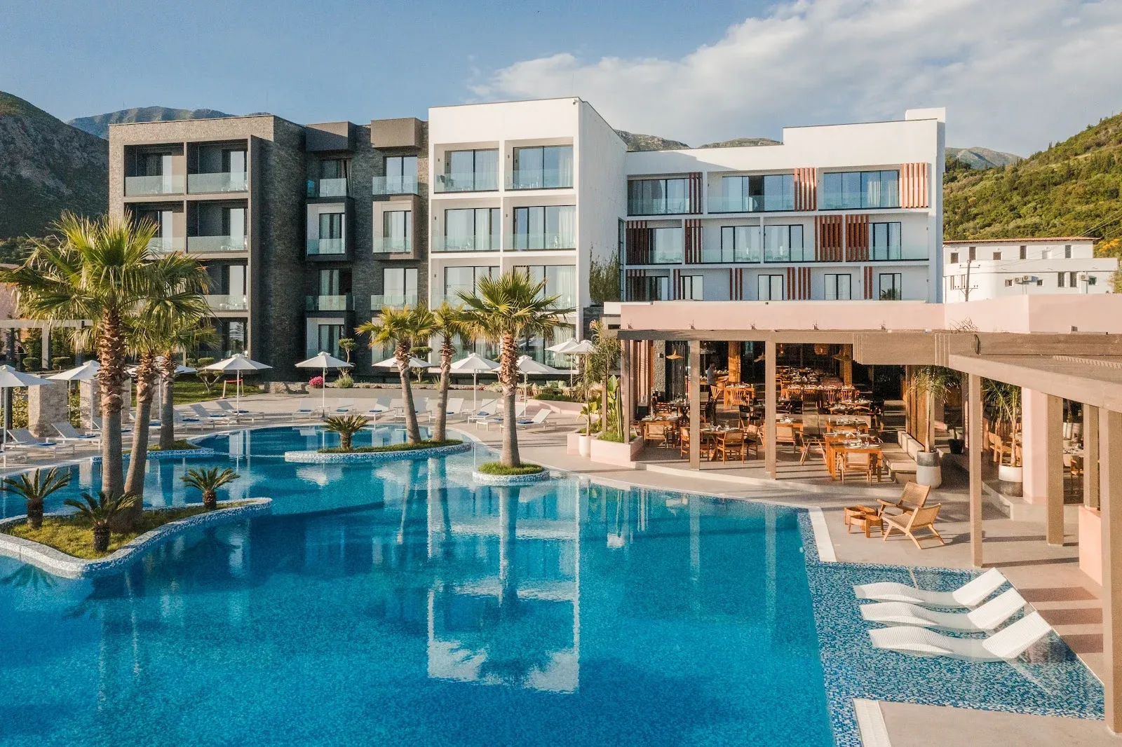 The white and blush façade of Miamar Luxury Hotel behind its outdoor pool, palms and restaurant terrace at Livadh, Himarë