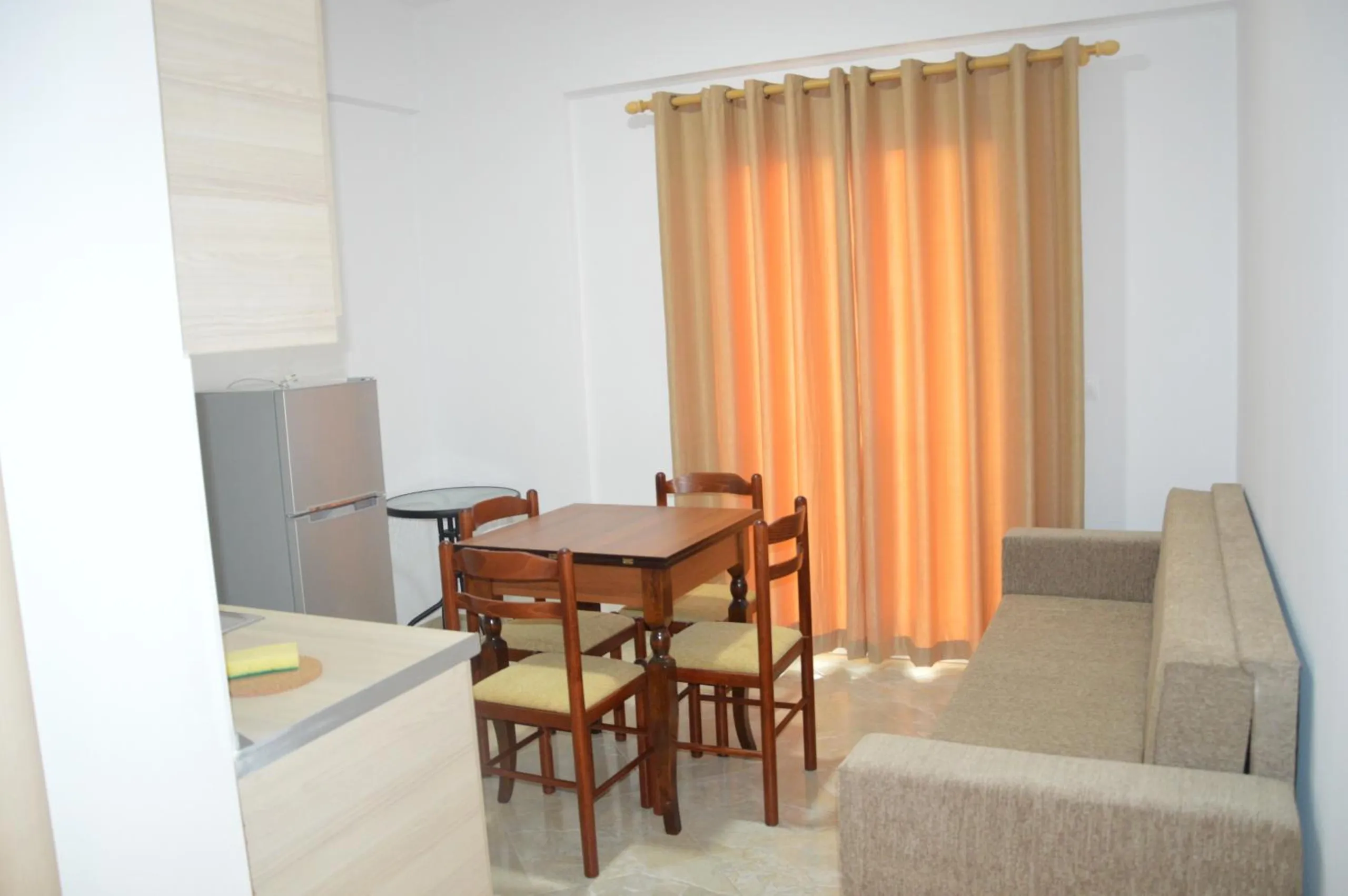 A compact kitchen with a fridge and a four-seat dining table, a sofa beside it and orange curtains at the window
