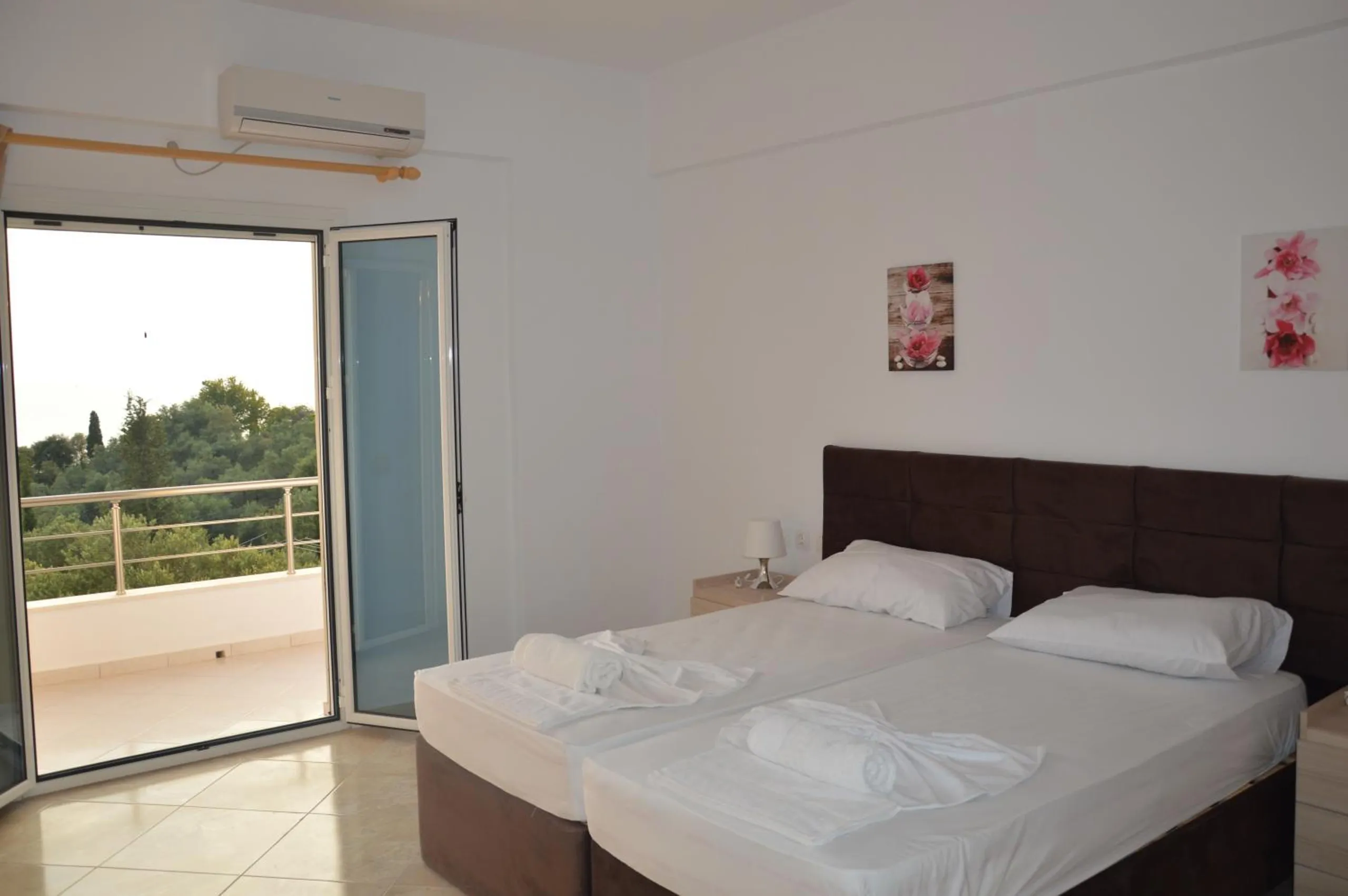 A bedroom with two beds pushed together under a dark headboard, white towels, and the balcony door open to green trees