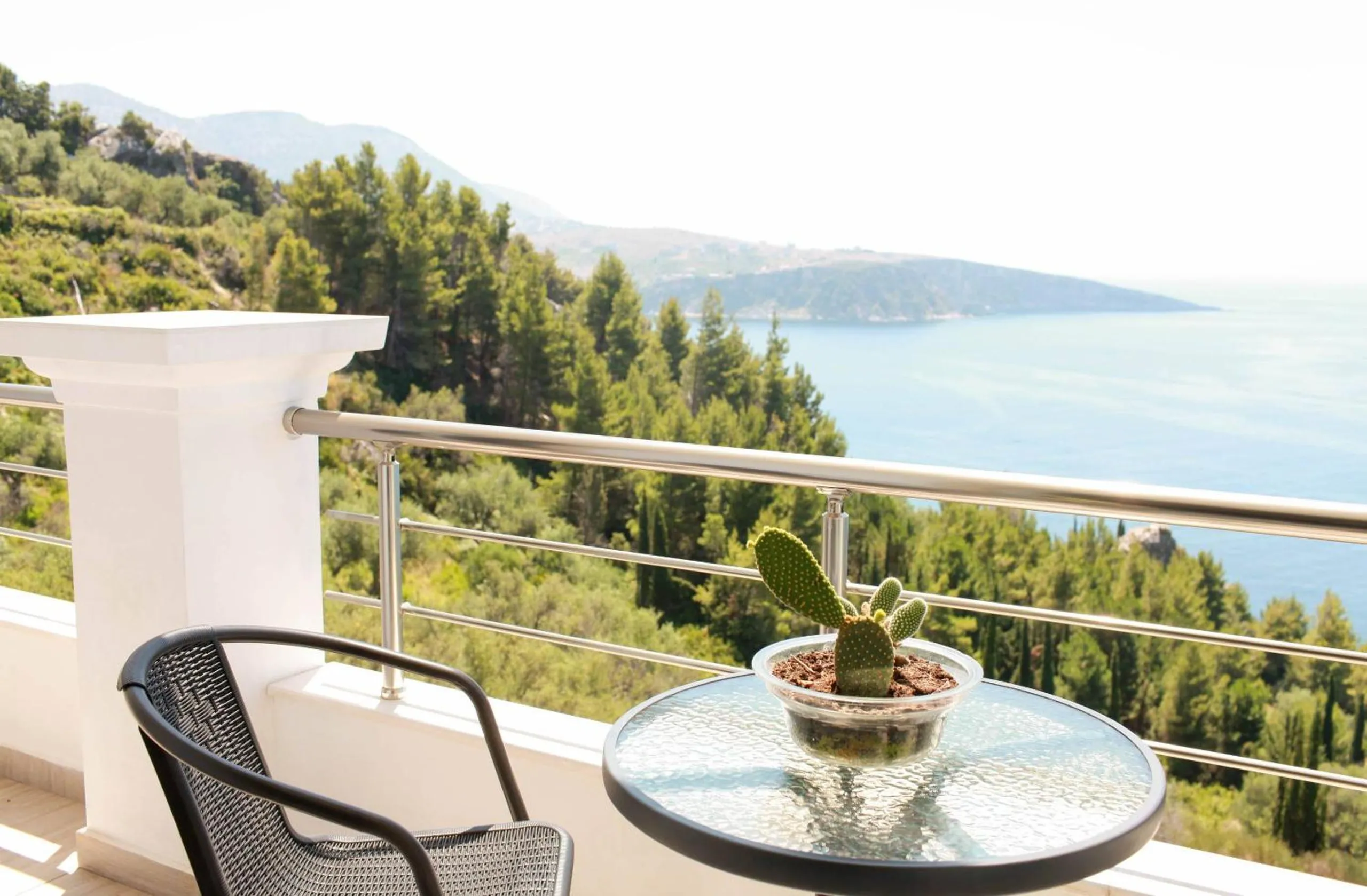 A glass balcony table with a small cactus in a bowl and two wicker chairs, above a wooded slope that runs down to the bay and a headland