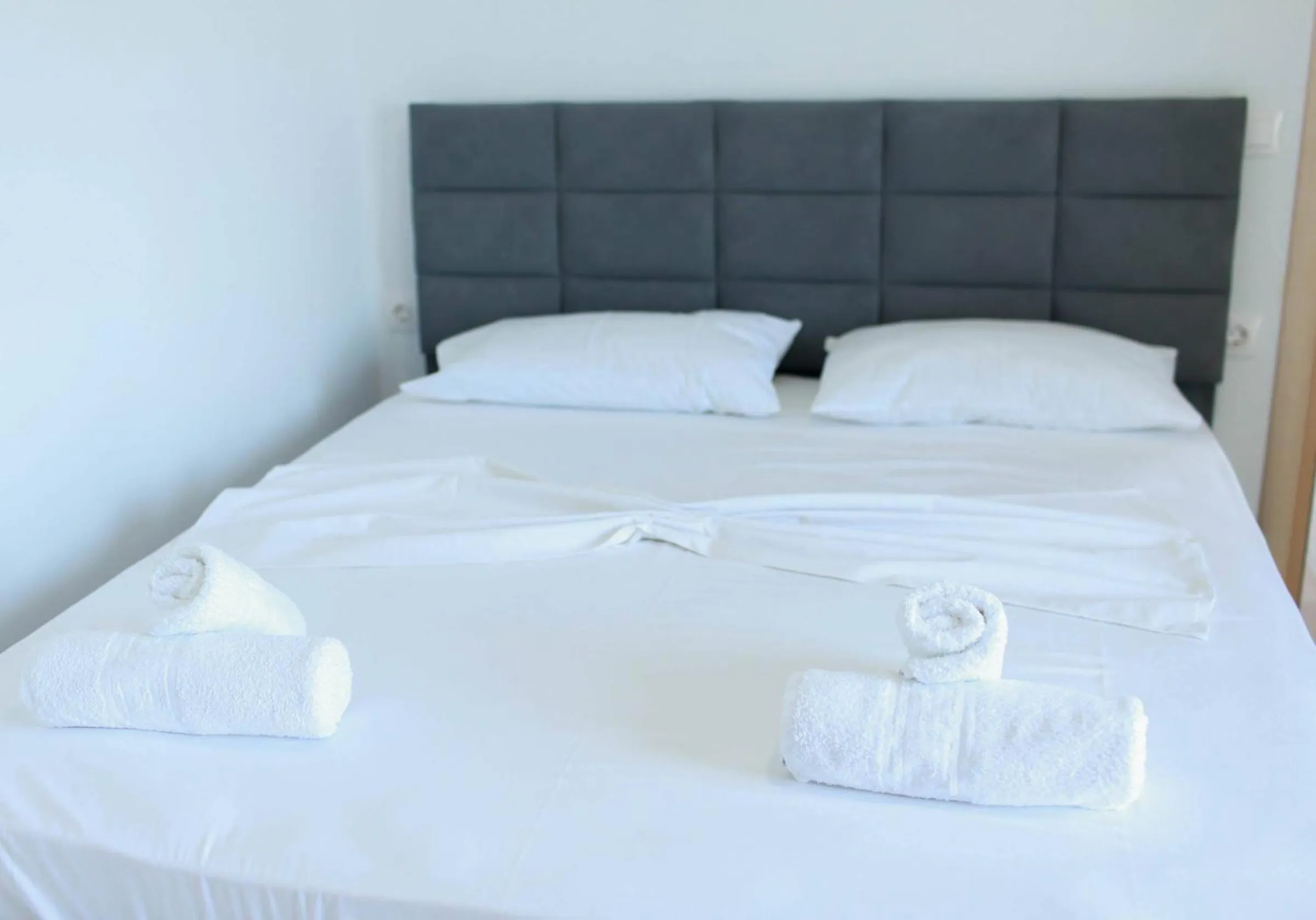 A double bed made in white with folded towels under a grey upholstered headboard