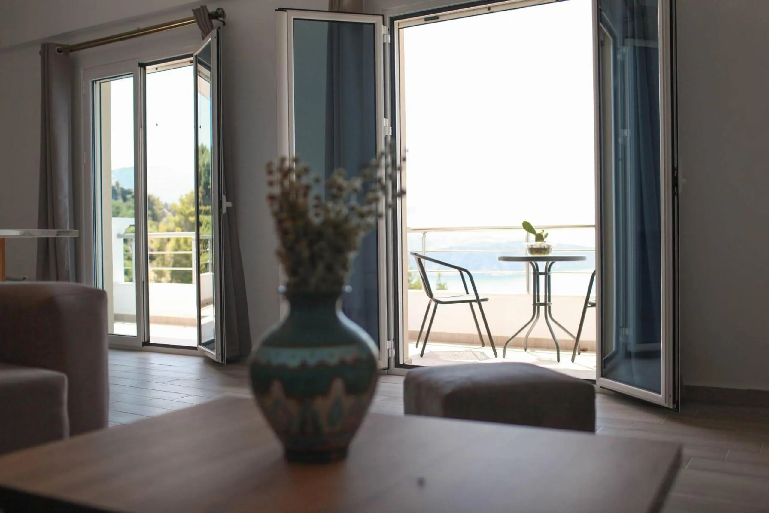 A Deluxe Apartment living room: a grey corner sofa, a table with a ceramic vase, and the balcony doors open on a table, two chairs and the sea