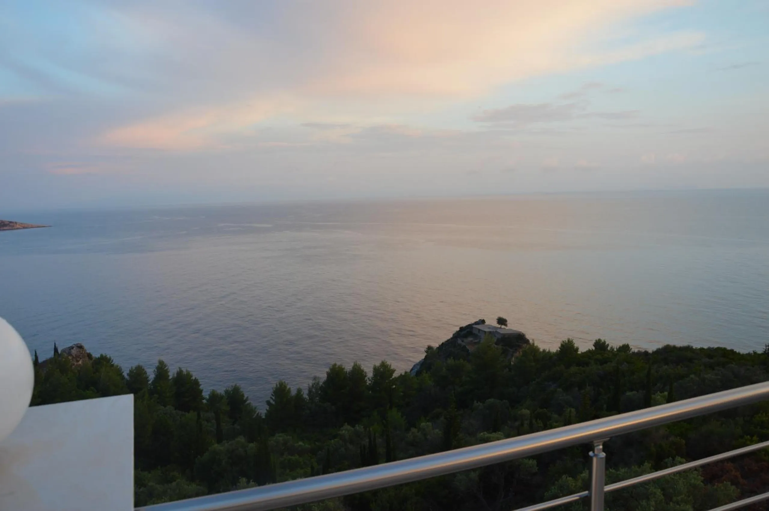 The sea at dusk from an upper balcony rail, a wooded slope below and a pale sky