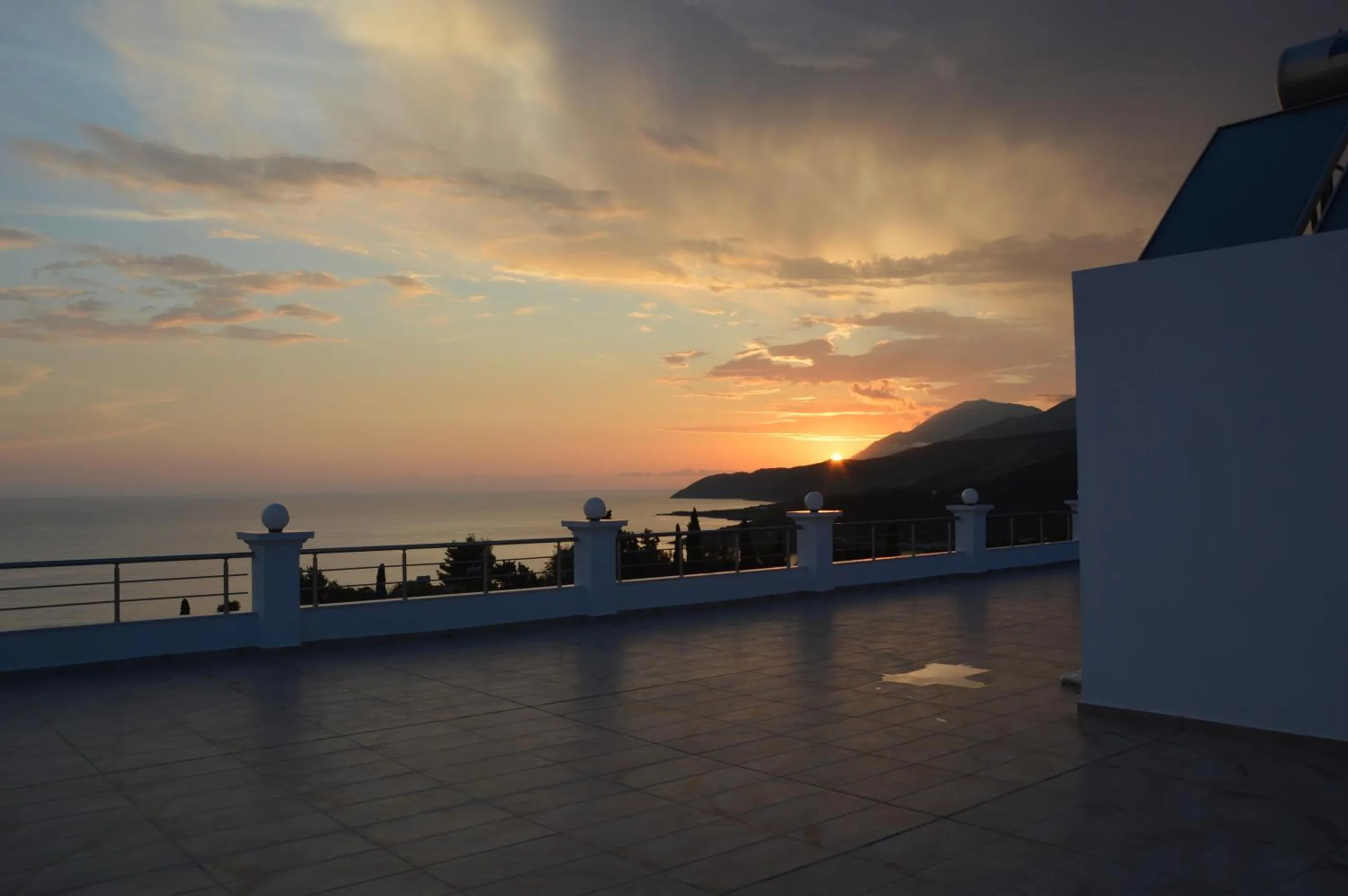 The tiled roof terrace of Mihalis Apartments at sundown, a white balustrade with globe lamps in front of the sea, the sun setting behind a mountain headland
