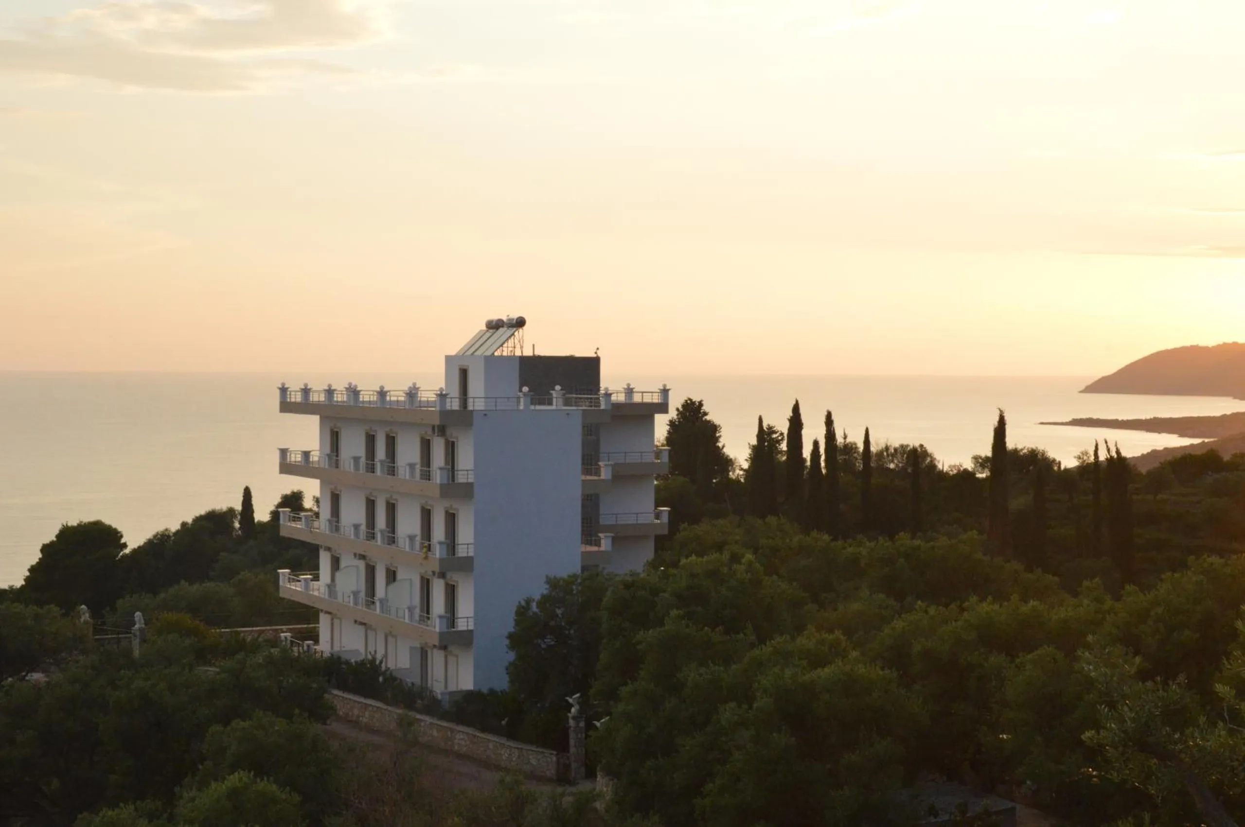 Mihalis Apartments at sundown: a white five-level block with balconies on every floor, standing in olive trees and cypresses on the hill above the Ionian, a headland on the right horizon