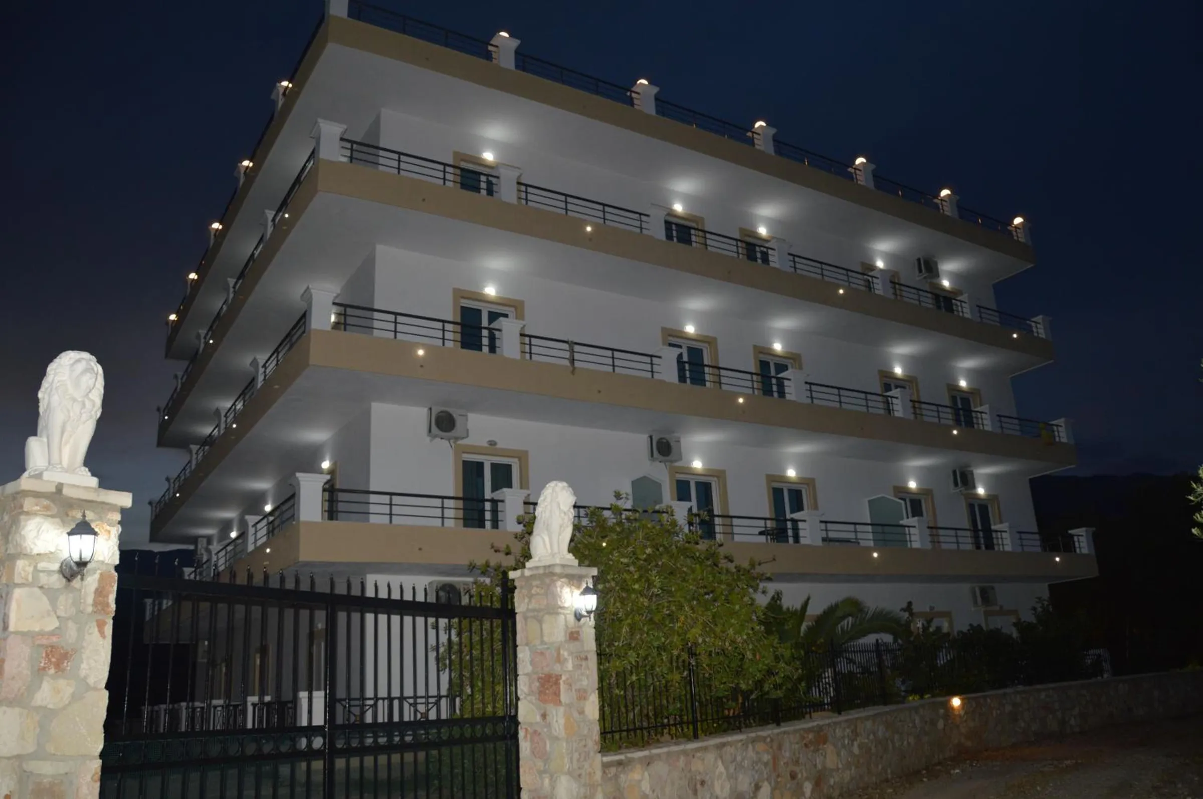 Mihalis Apartments at night, a light under every balcony slab, seen past a stone gatepost topped with a white statue