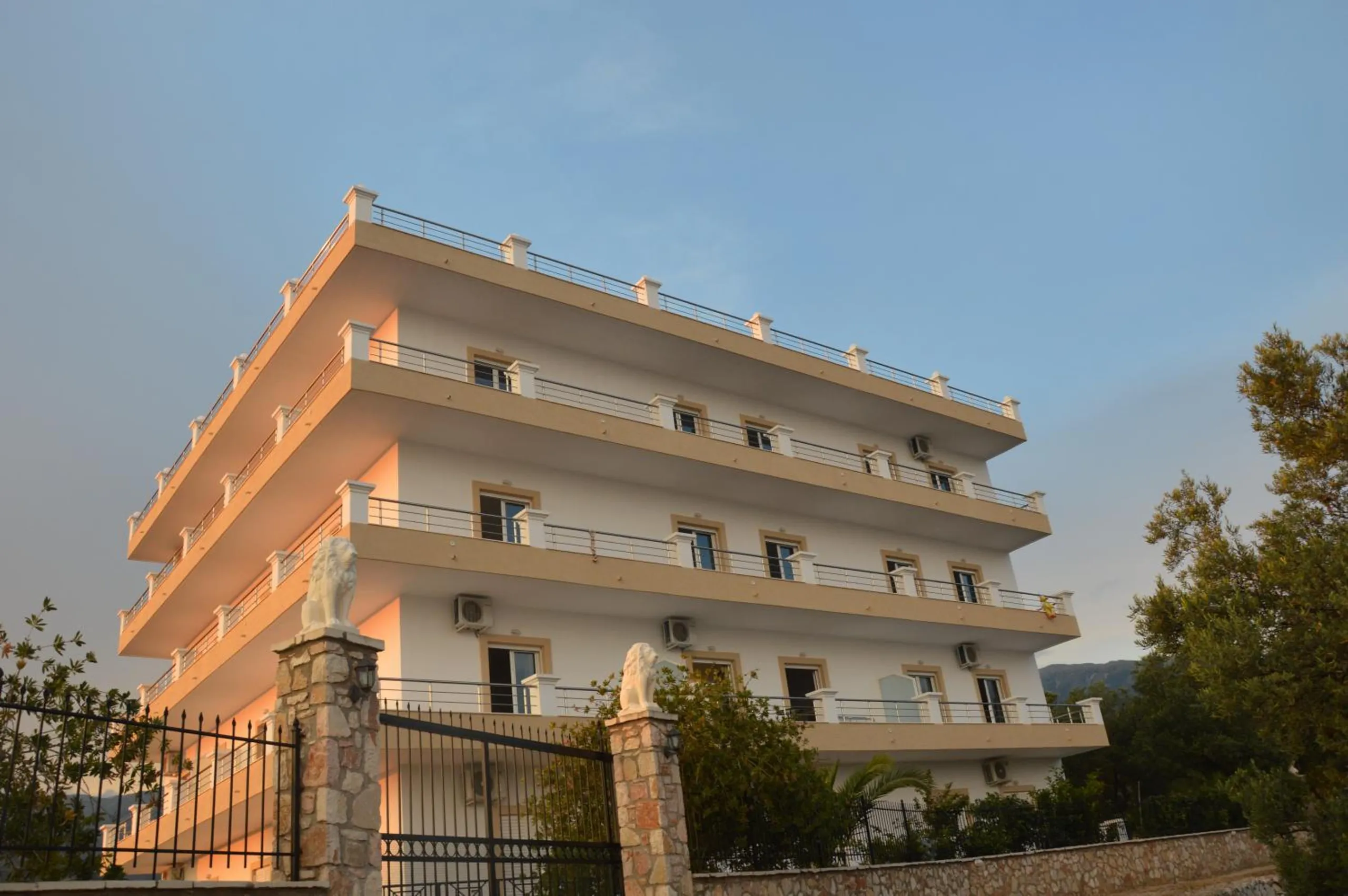 The front of Mihalis Apartments in evening light: four balcony storeys with sand-coloured window surrounds, behind an iron gate whose stone posts each carry a white lion