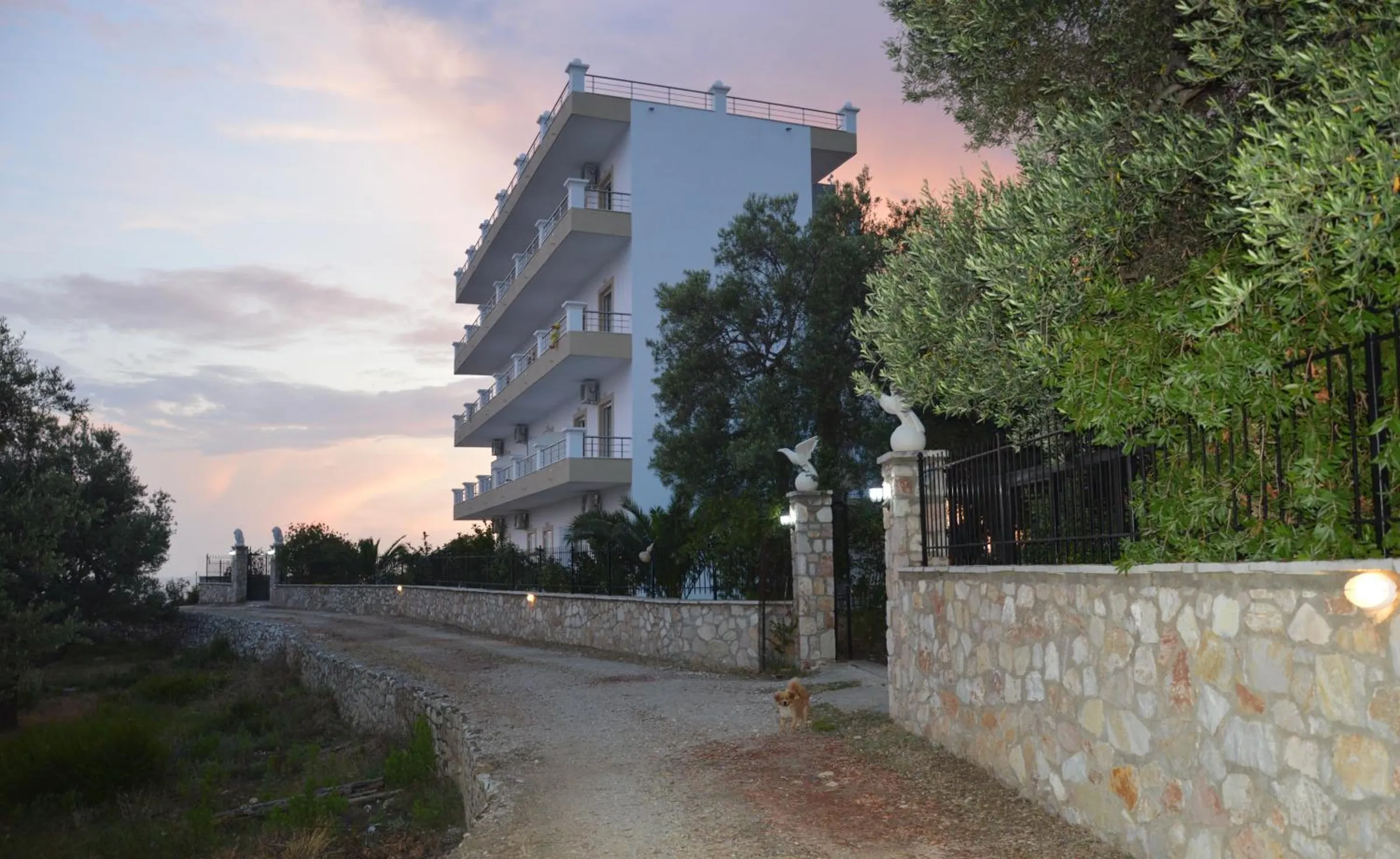 A gravel track running between dry-stone walls to the side gate of Mihalis Apartments at dusk, white bird statues on the gateposts and the block rising behind them