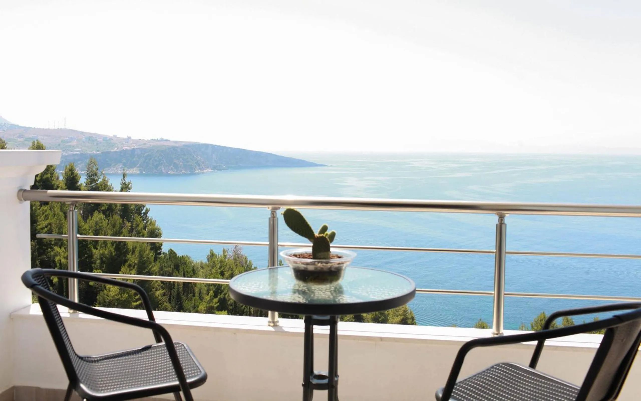Two black wicker chairs and a glass table on a balcony, facing a calm sea with a low headland on the left