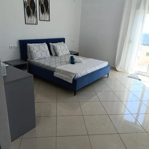 A blue upholstered double bed under two framed leaf prints in an apartment at Nikolari Resorts, cream floor tiles running to a curtained balcony door