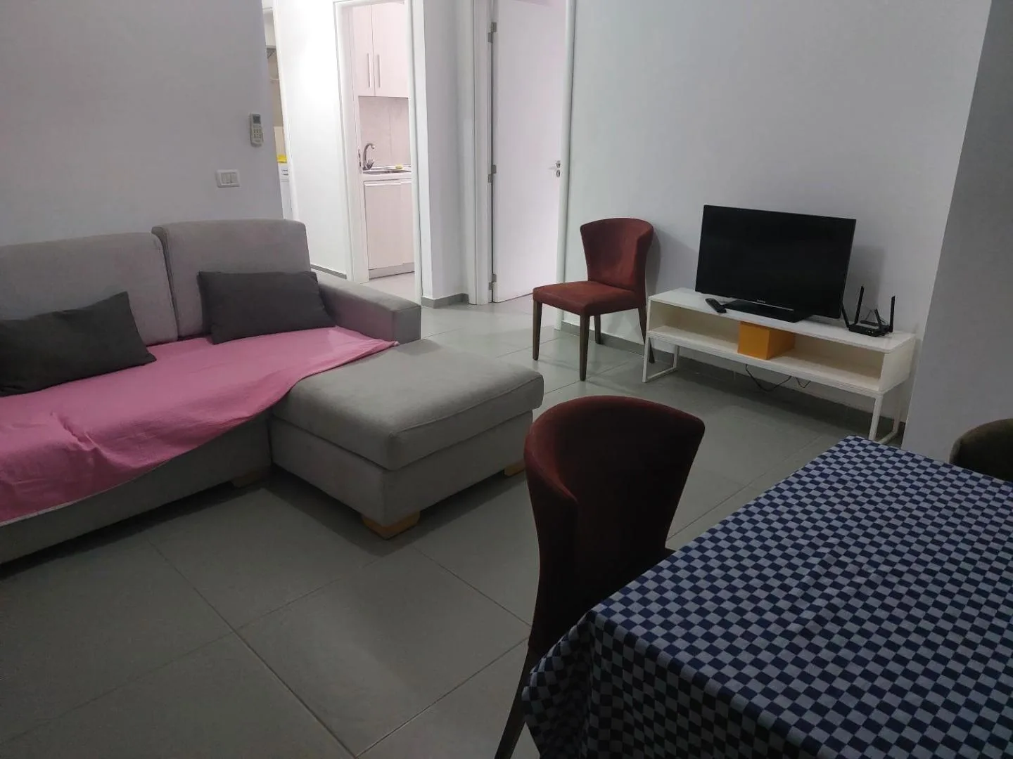 The living room of a two-bedroom apartment: a grey corner sofa, two red chairs, a television and a checked tablecloth