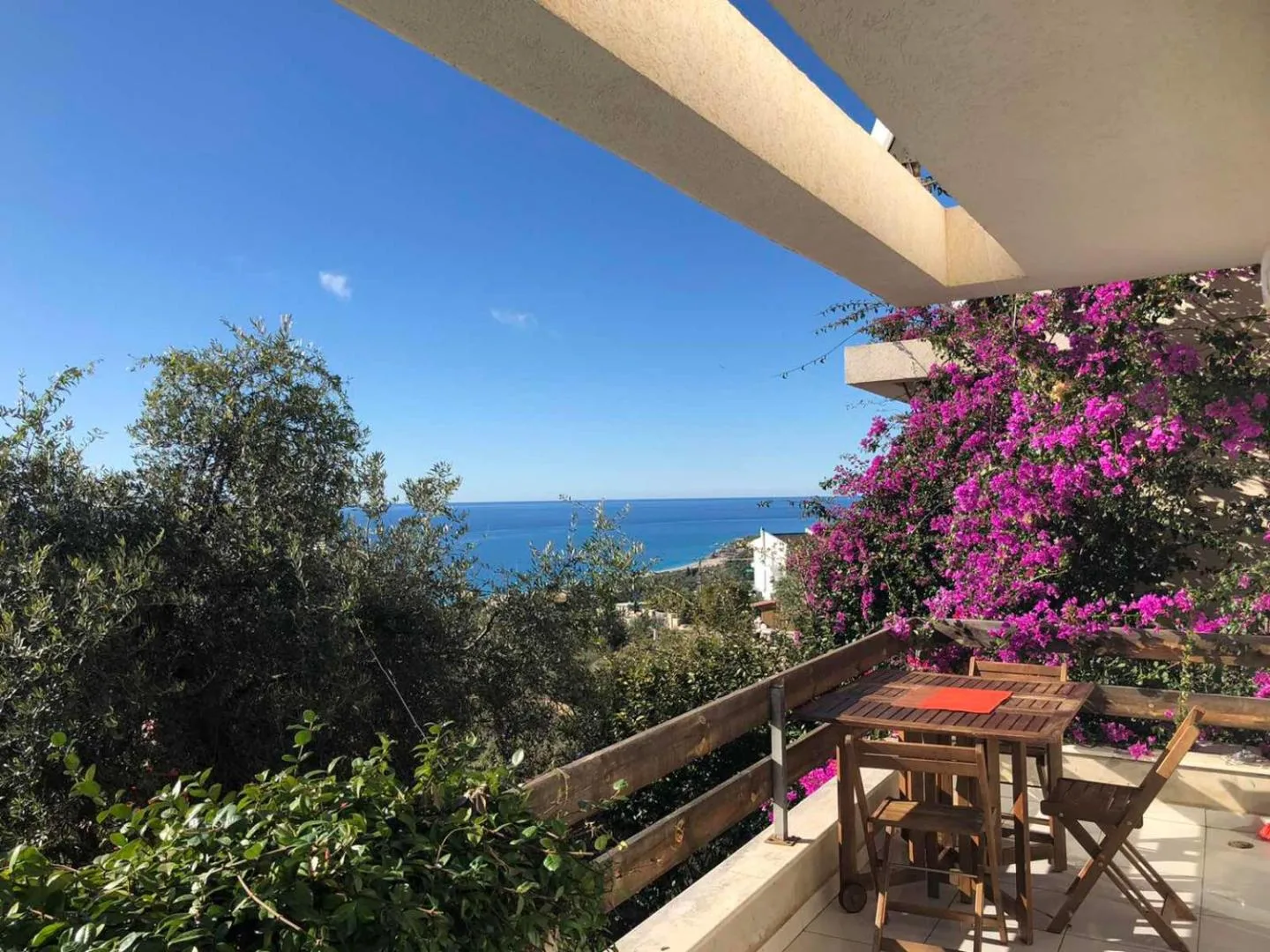 A terrace with a small wooden table under bougainvillea, looking over olive trees to the sea