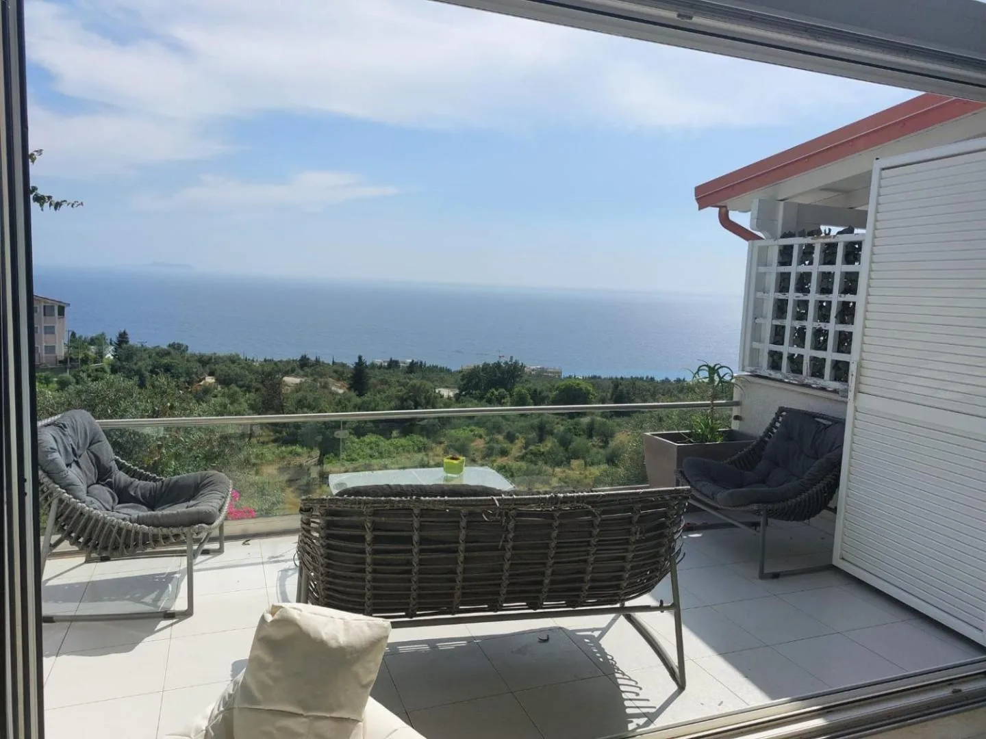 A balcony with grey woven armchairs, looking down over the hillside to the sea