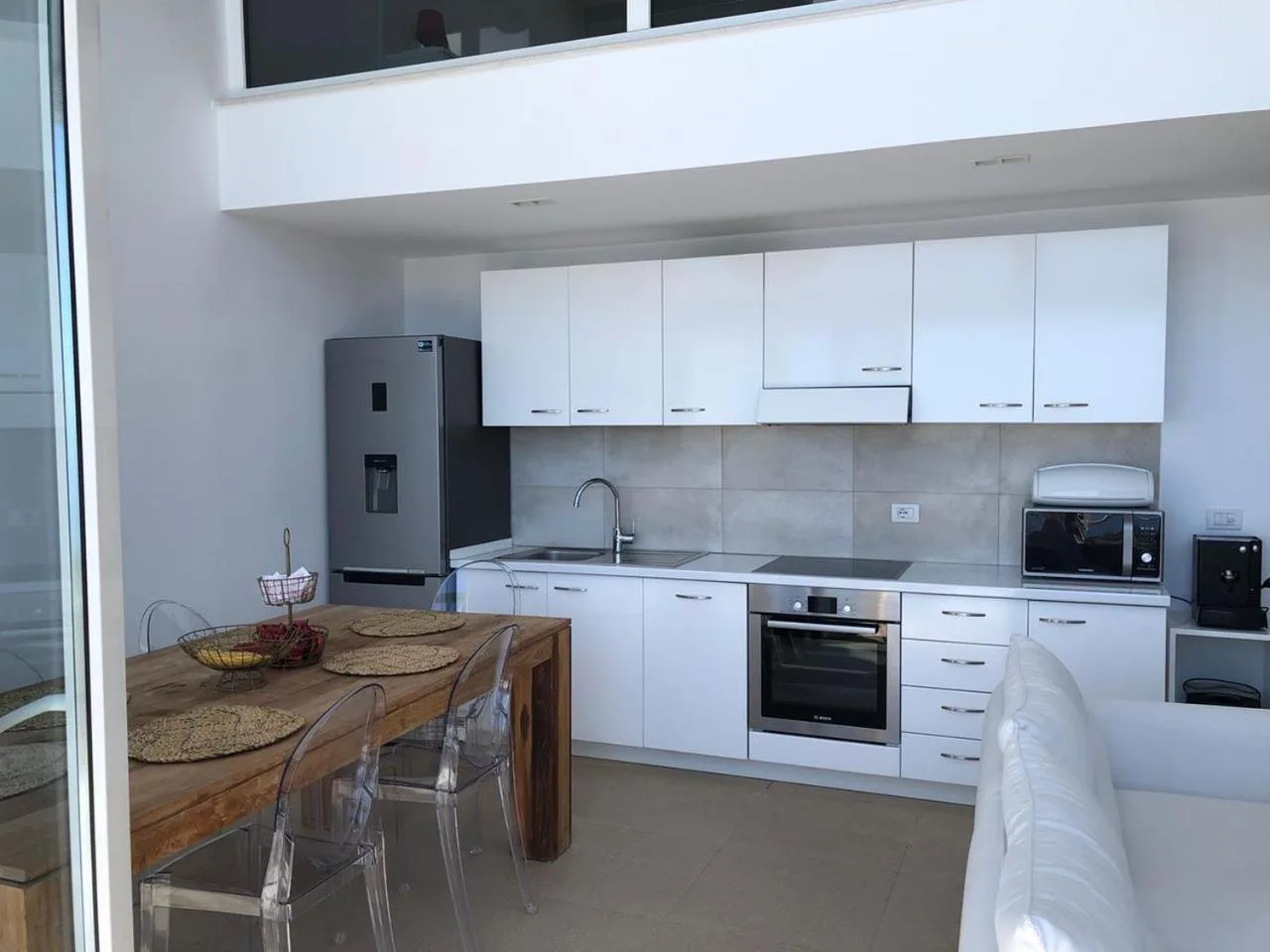 A white kitchen with a wooden dining table, under a double-height ceiling