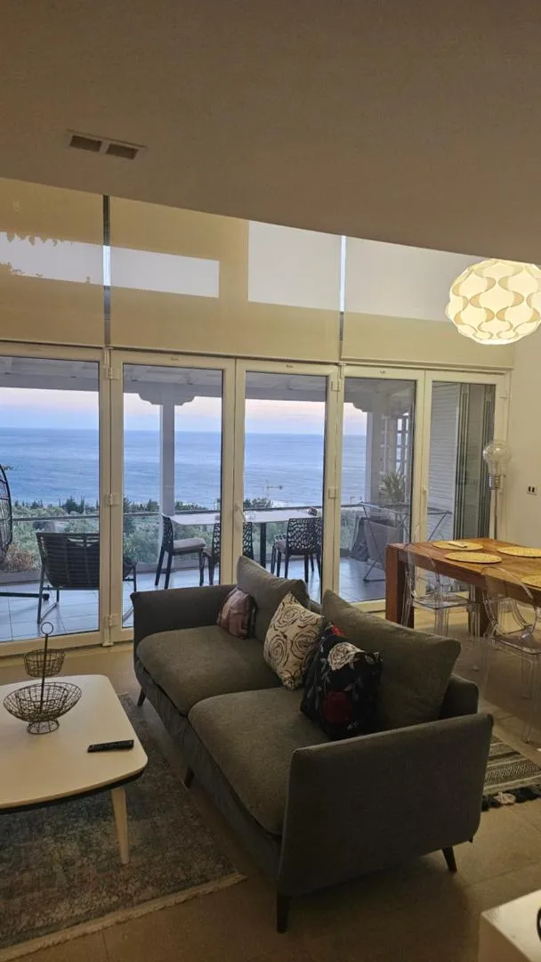 A living room with a grey sofa and full-height glazing onto a terrace and the evening sea