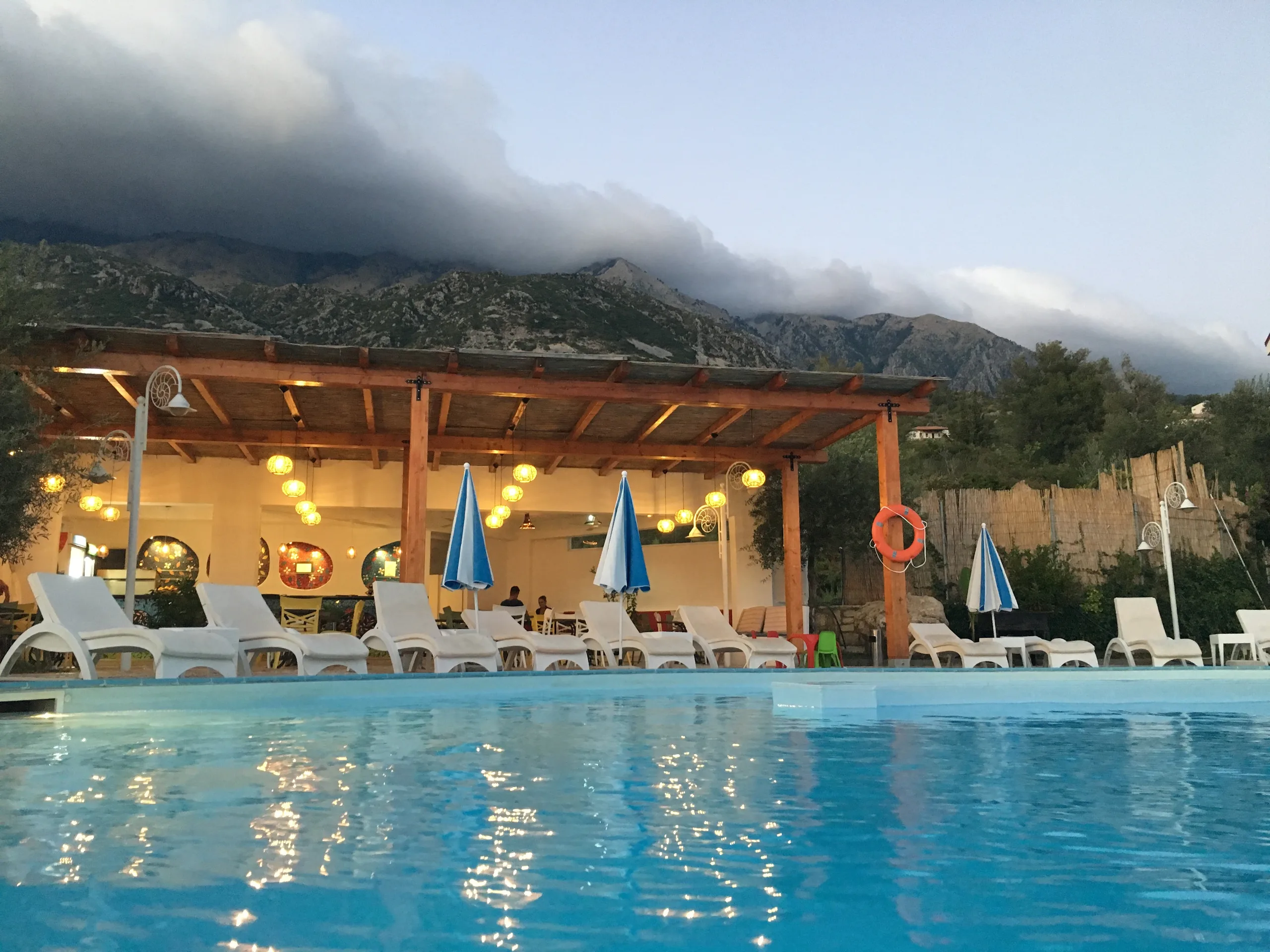 The pool and its timber bar at dusk under the Ceraunian mountains, with cloud resting on the ridge