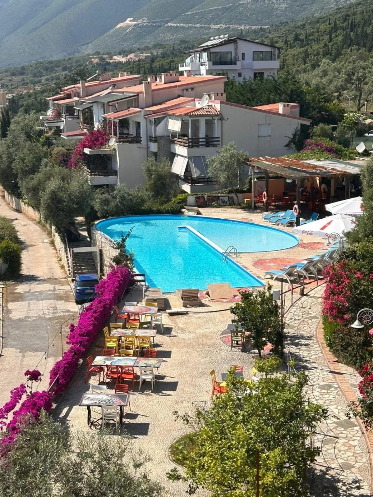 The pool at Olives Residence seen from above, with white villas and terracotta roofs behind it, olive trees, a bougainvillea hedge and the mountain beyond