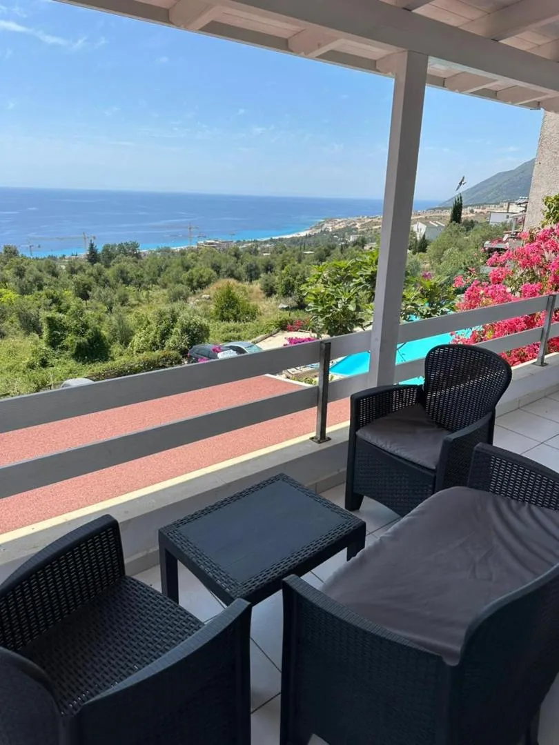 A balcony with dark wicker chairs looking over olive trees and the pool to the coast