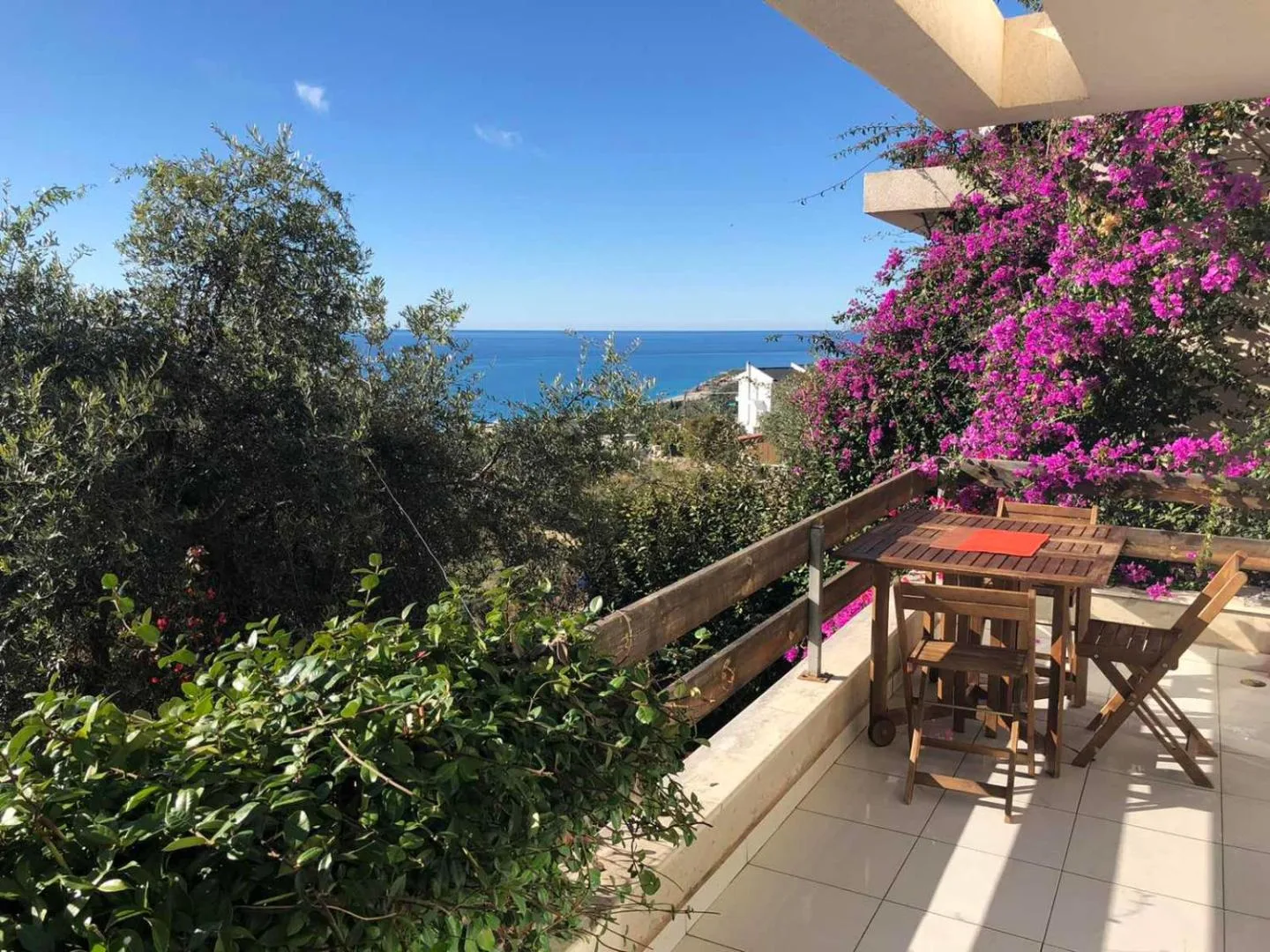 A tiled terrace with a wooden table and chairs under magenta bougainvillea, looking over olive trees to the sea