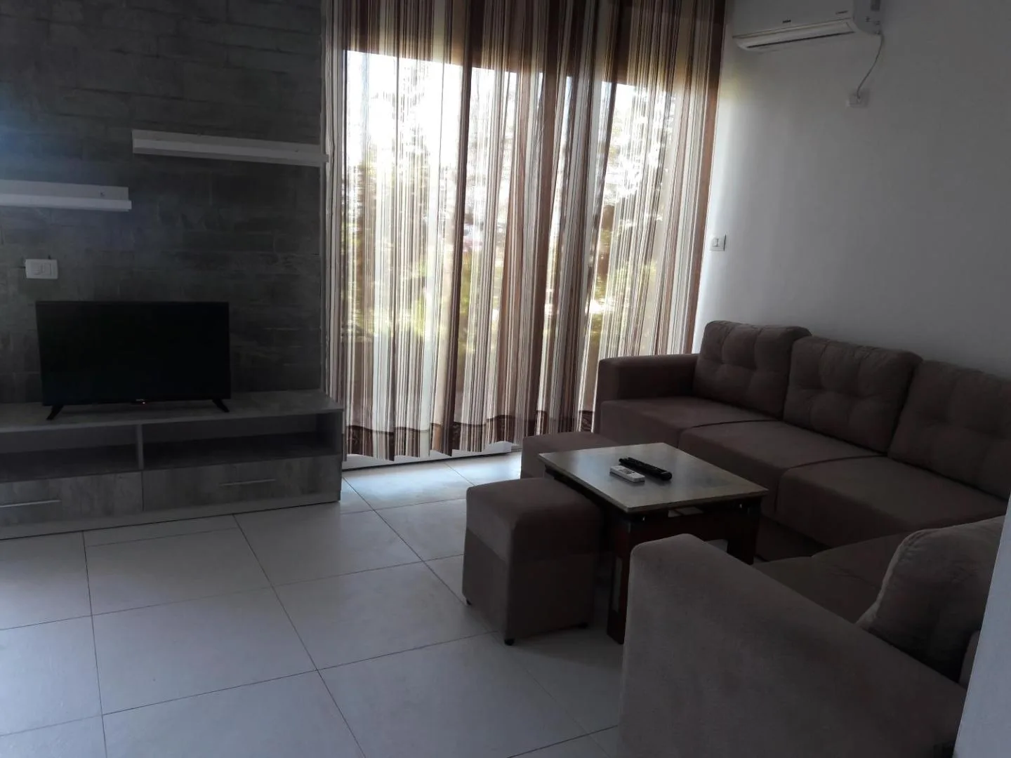 A villa living room with a brown corner sofa, a television and long curtains over glass doors
