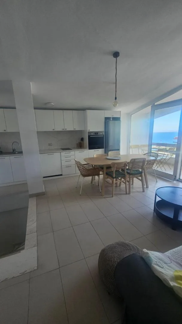 A white kitchen with an oven and a wooden table, a window looking out to the sea