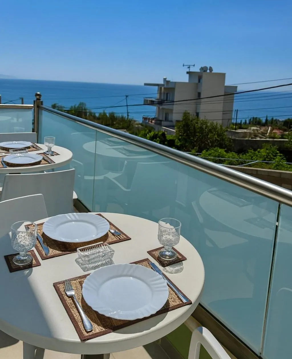 A round balcony table laid for two with plates and crystal glasses, the glass balustrade and the sea beyond it