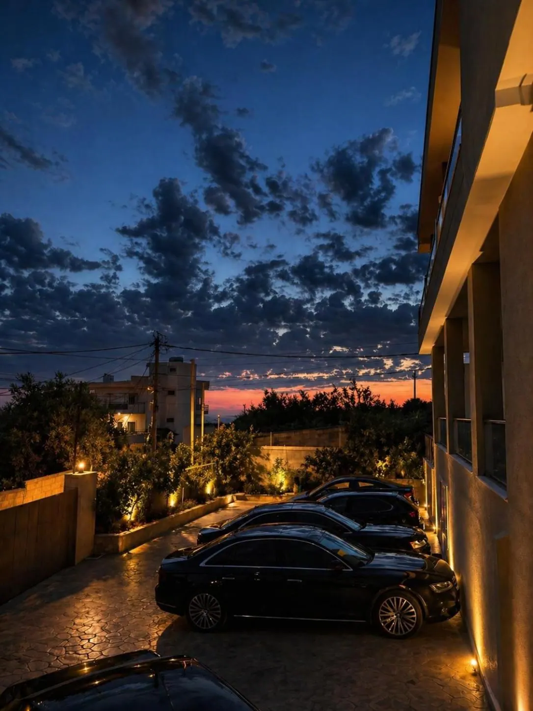 The courtyard after sunset, cars parked on stamped paving beside the house, low garden lights along the wall and a red band of light over the sea