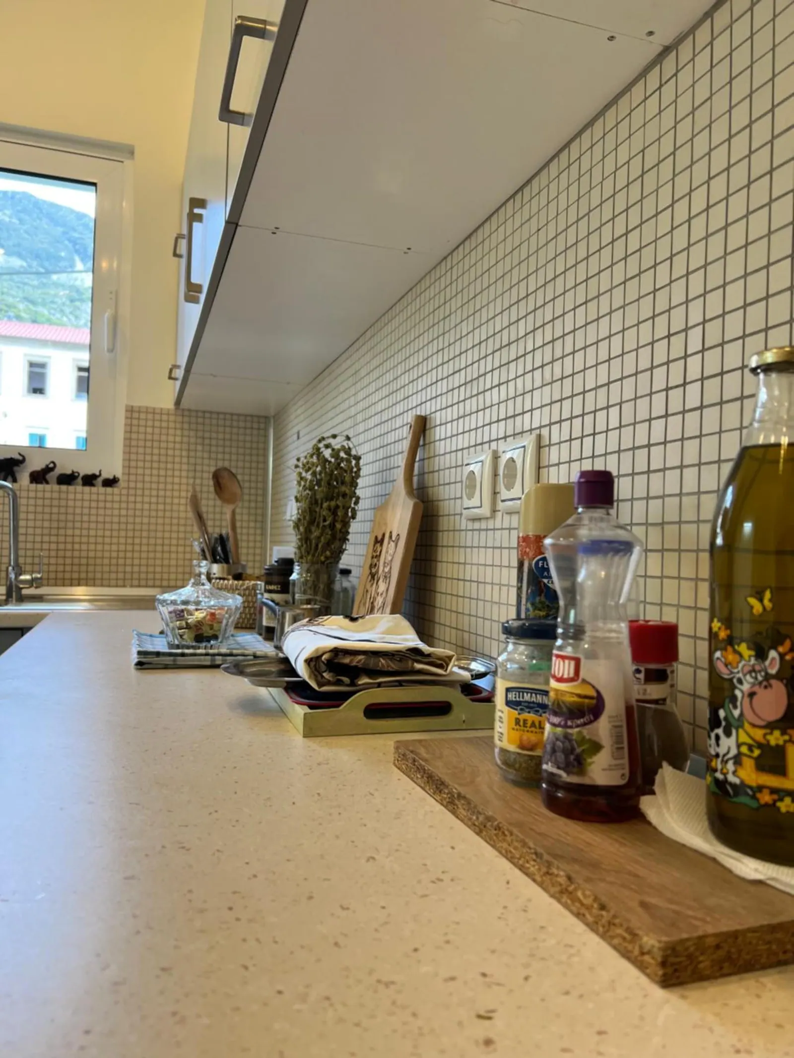 A long kitchen counter under white tiles with a bottle of olive oil, oils and spices, a sprig of herbs and a wooden board by the window