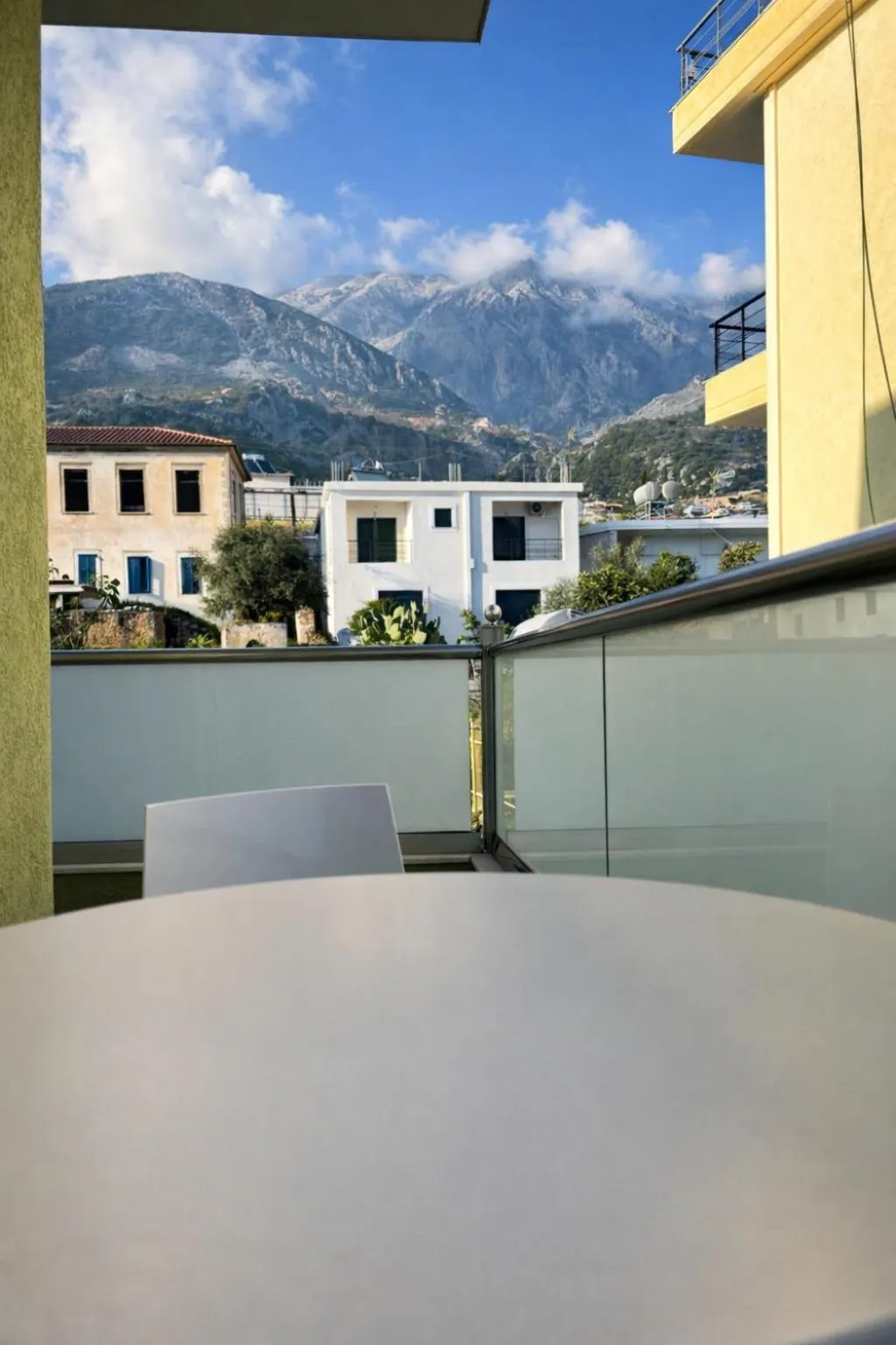 A balcony table under a pale yellow wall, with the white houses of the village and the snow-dusted Ceraunian mountains behind