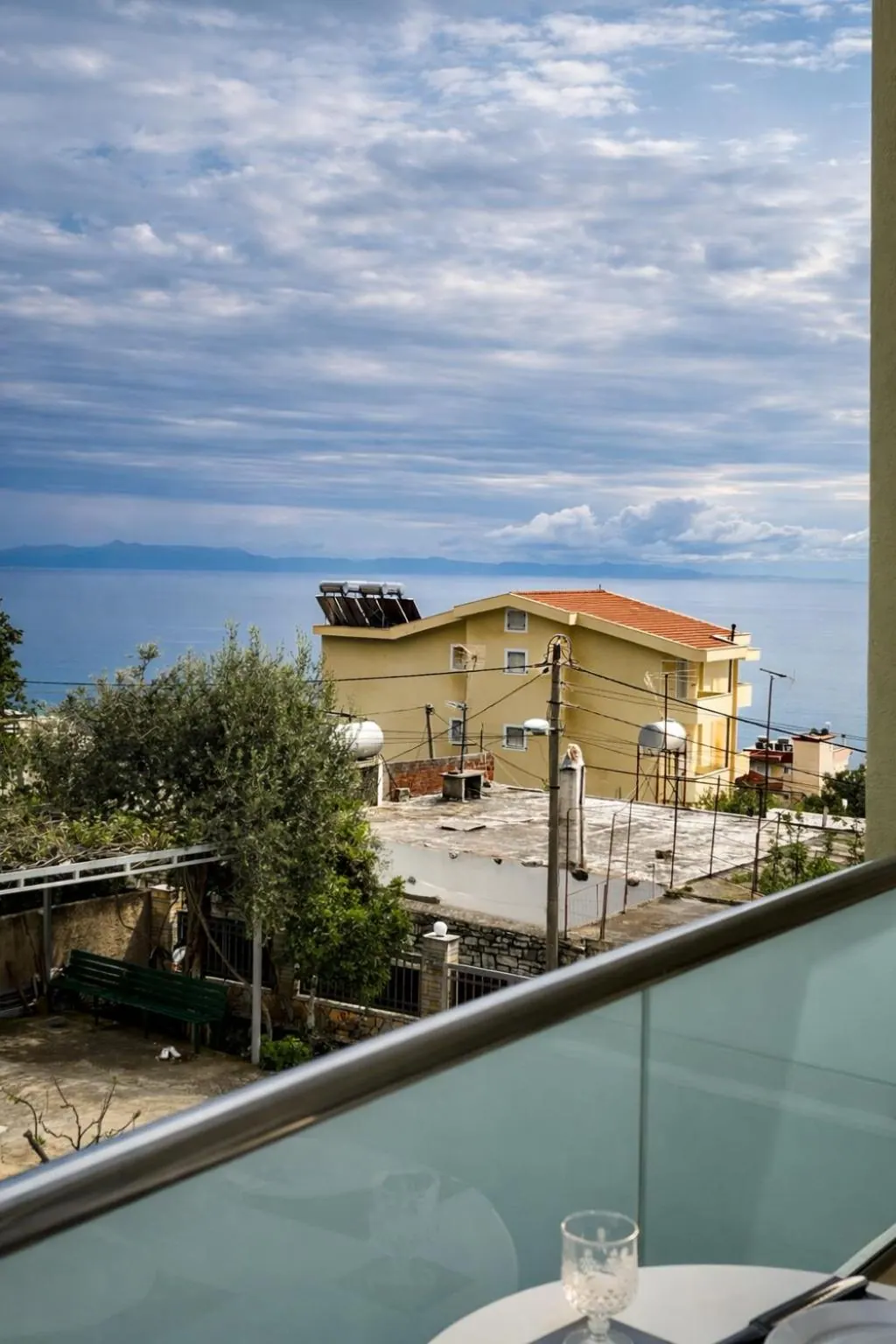 Over the balcony rail: olive trees, flat roofs and a yellow house with a red-tiled roof, and the sea and the far coast under a broken sky