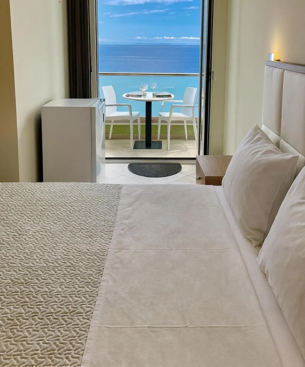A white double bed, and beyond it an open door to a balcony with a round white table and two chairs and the open sea behind the glass